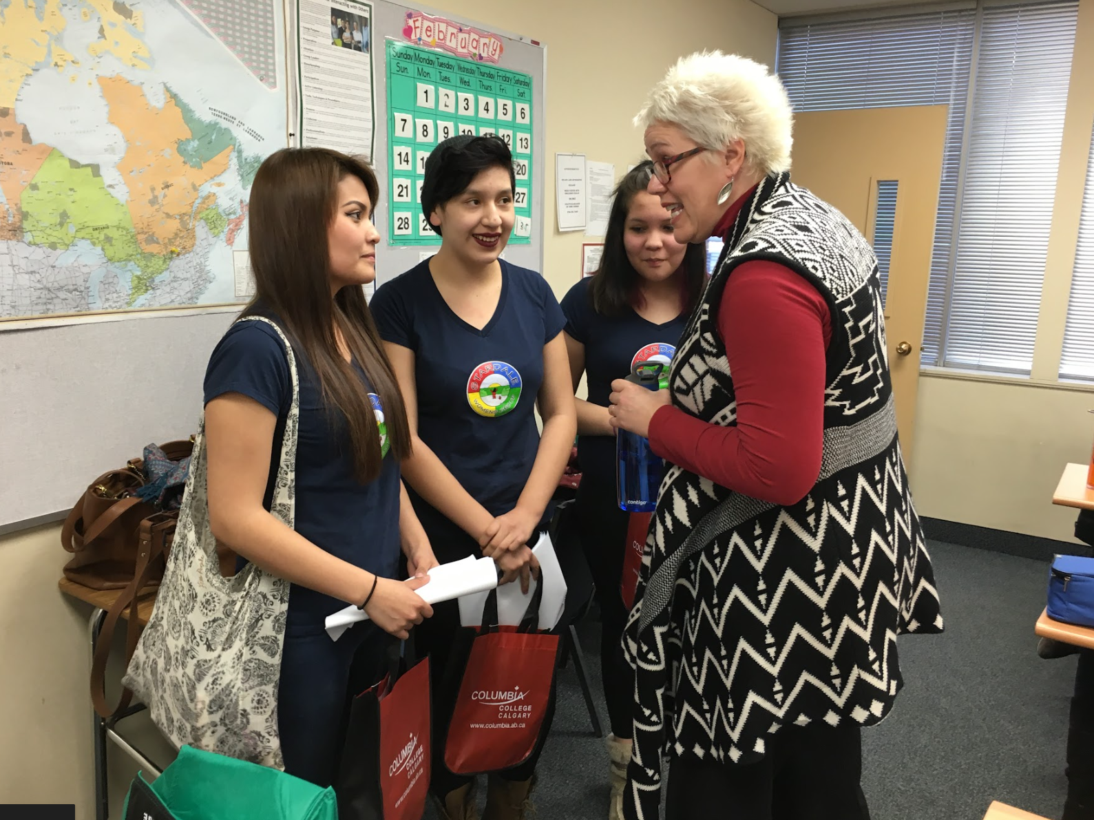 Four women, three young students and one older woman, engaged in conversation in a classroom. The students are wearing navy blue shirts with a rainbow and university logo. The older woman is talking to them, holding a blue folder. Behind them is a large map of North America and a February calendar on the wall.