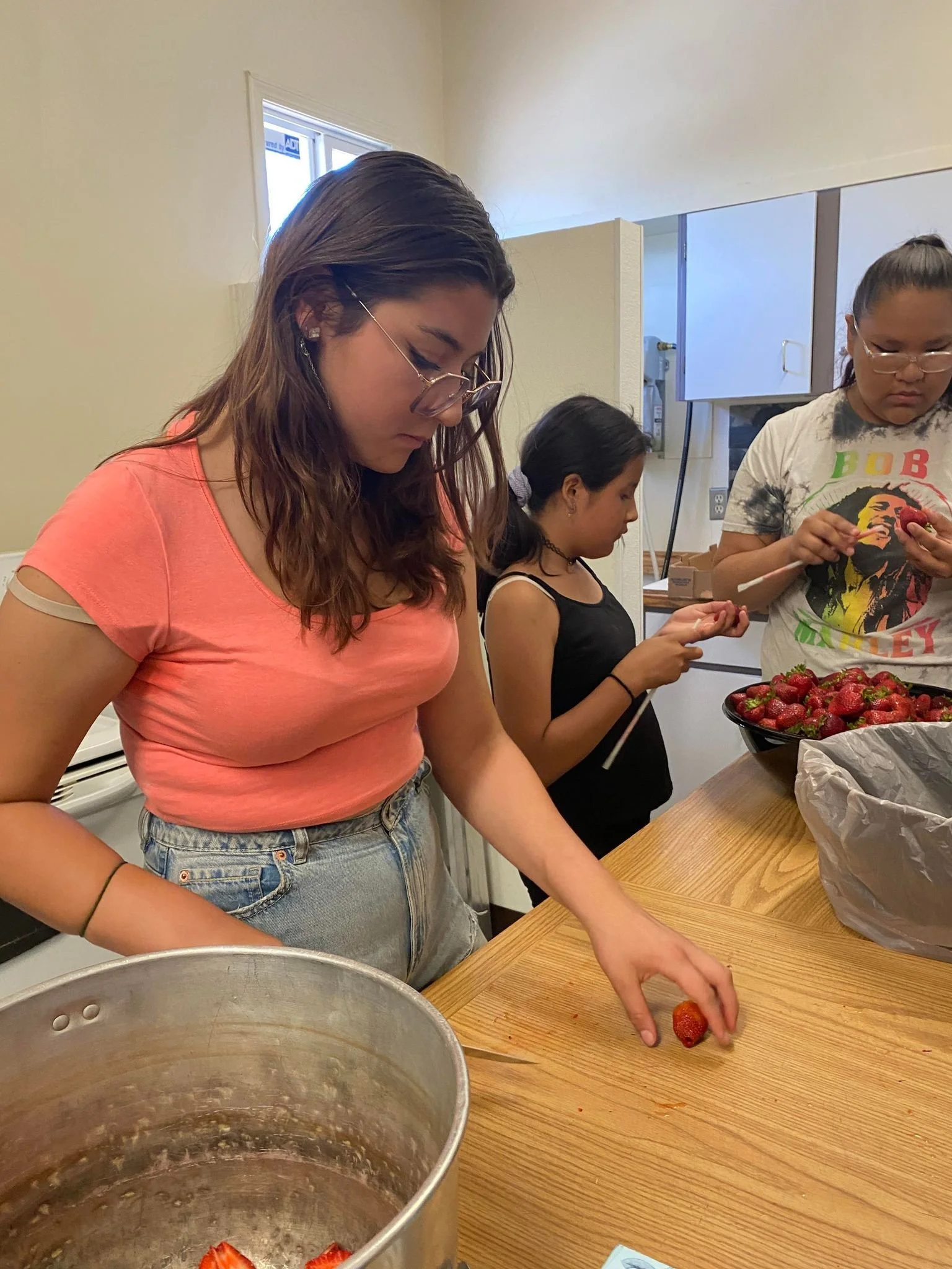 Three girls preparing strawberries in a kitchen, with one girl in the foreground, and two girls in the background also working with strawberries.