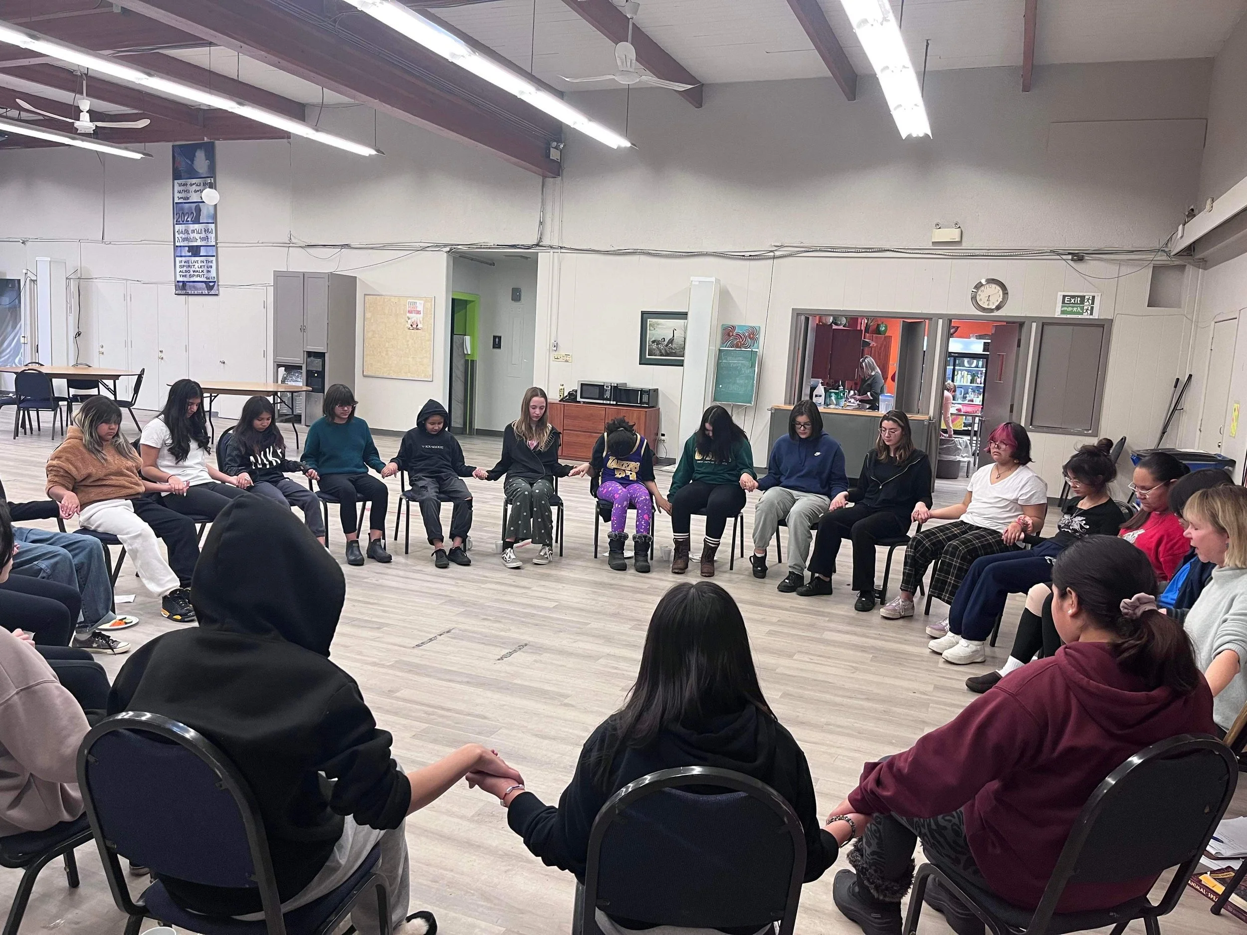 Group of women and girls holding hands, sitting in a circle in a community room.