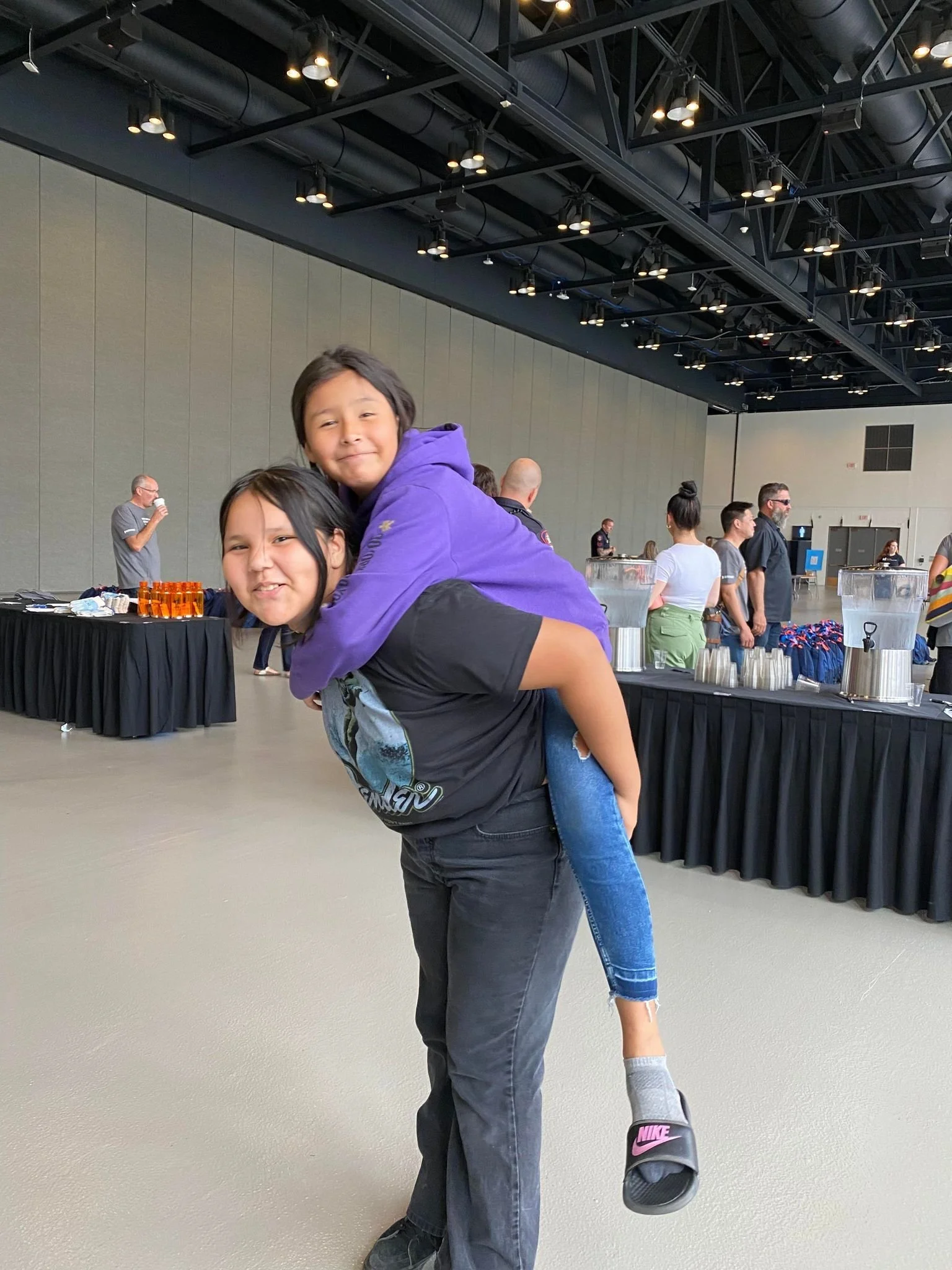 Two children, one giving the other a piggyback ride inside a large indoor venue with high ceilings and a food or drink station in the background.