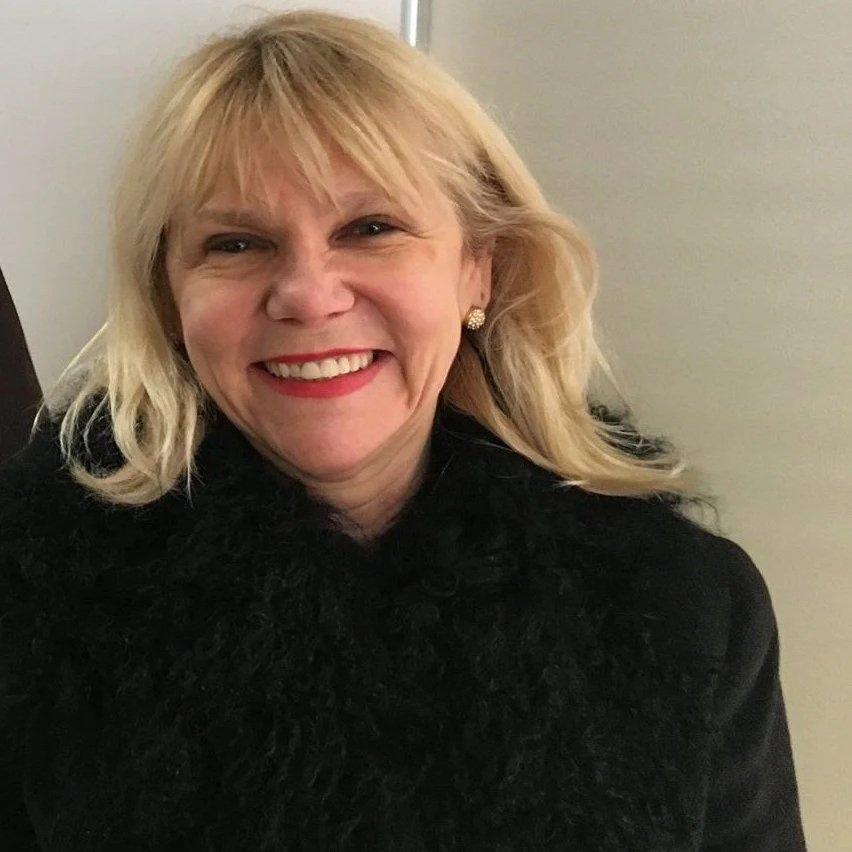 A smiling woman with blonde hair, wearing red lipstick, earrings, and a black coat with a furry collar, standing against a neutral background.