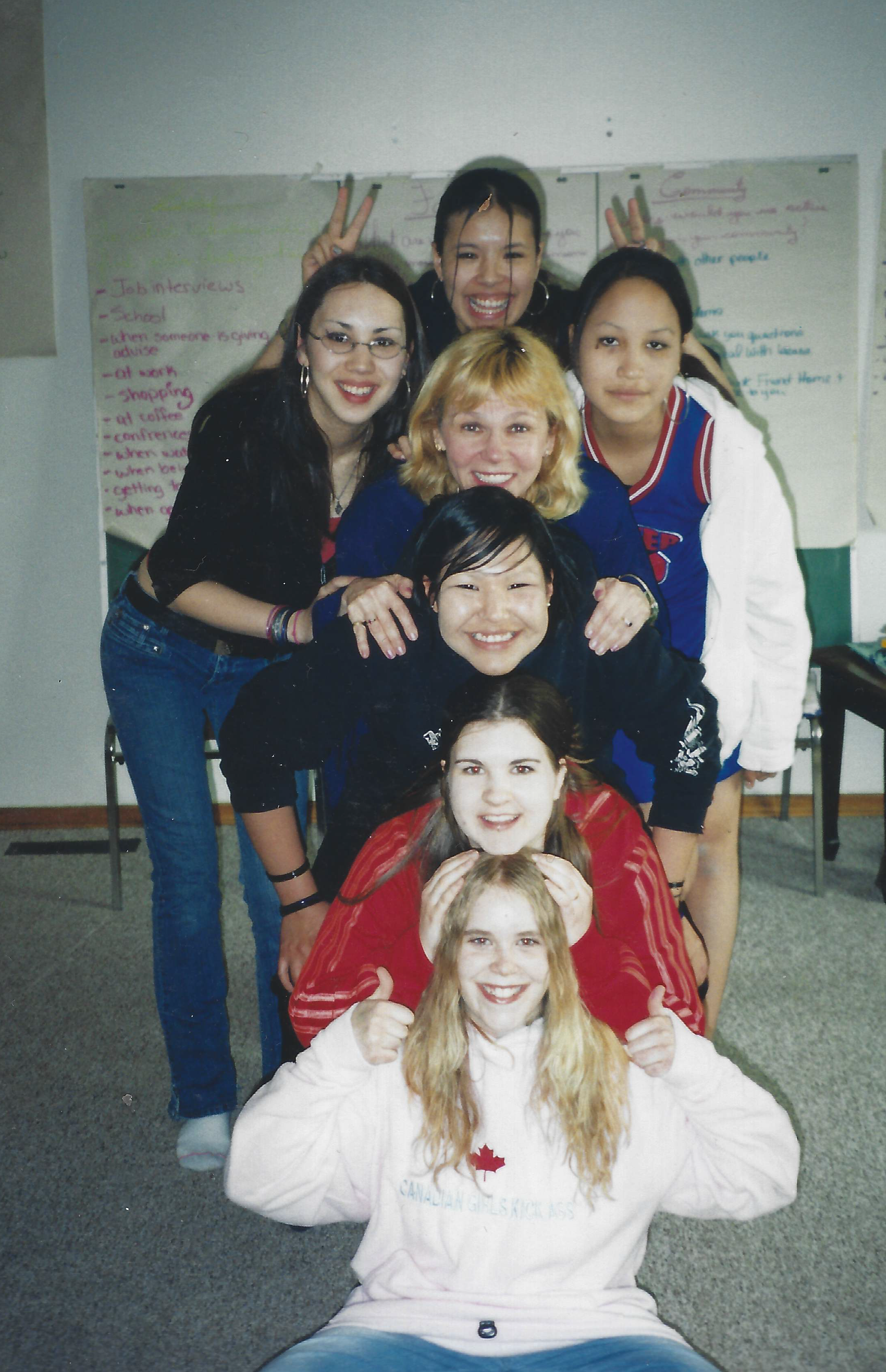 A group of nine young women stacking on each other in a classroom, smiling and making playful gestures.