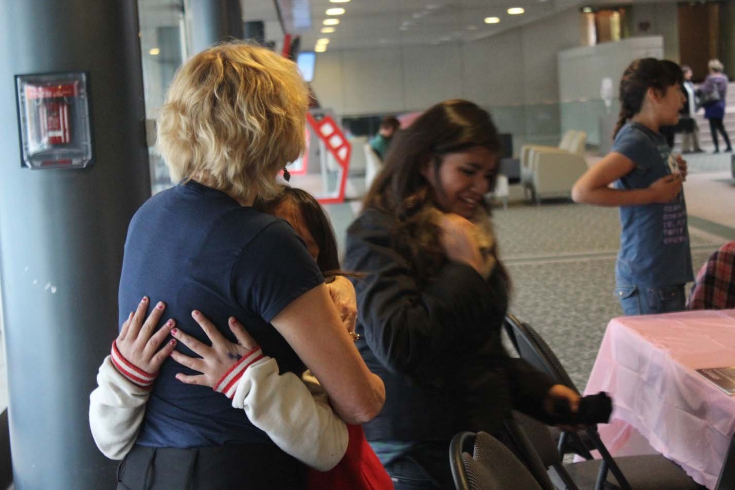A woman with blonde, curly hair embraces a young girl with her arms around the woman. A woman with long, dark hair in a black jacket is smiling nearby. In the background, a girl in a blue shirt stands, and other people are seated or walking in a wait