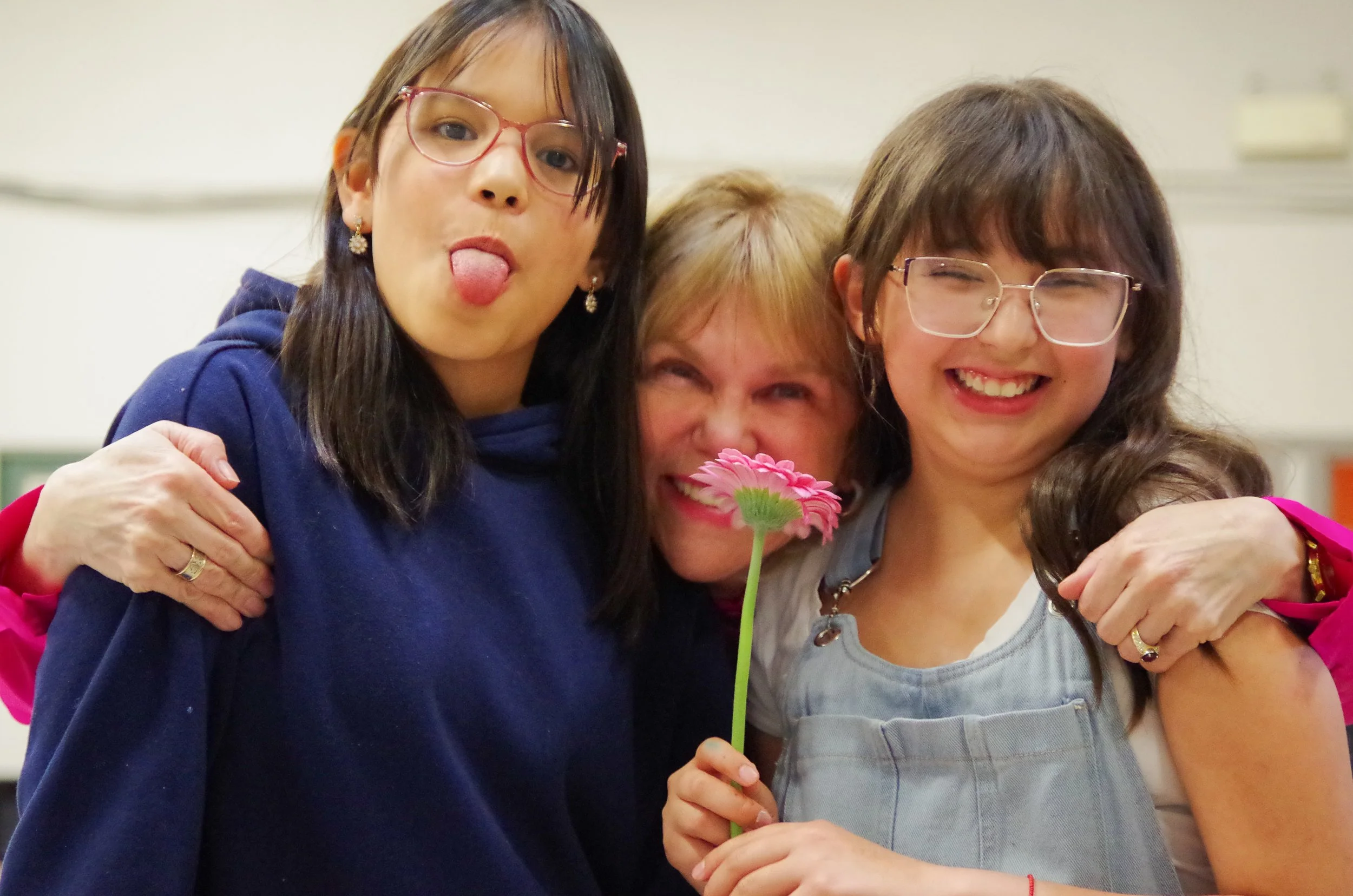 Three women smiling and hugging, one holding a pink flower in front of her face, in an indoor setting.