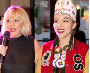 Two women standing side by side, smiling. The woman on the left has blonde hair and is holding a microphone, wearing a black top. The woman on the right has dark hair, is wearing traditional attire with a headdress and jewelry.