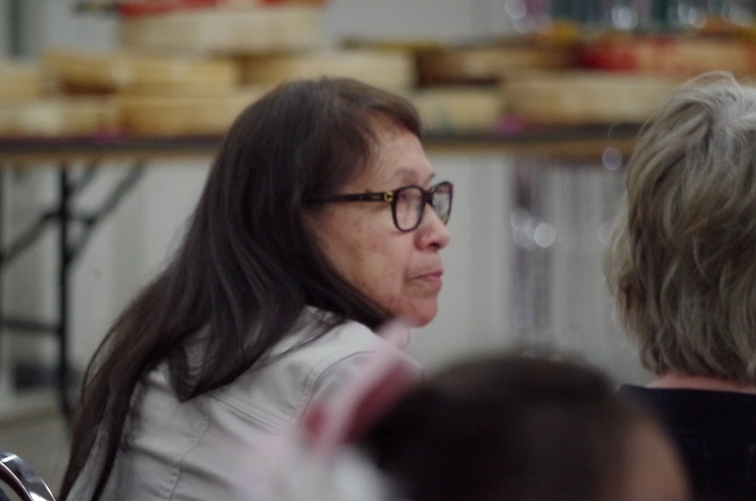 A middle-aged woman with long dark hair, glasses, and wearing a white shirt, sitting in a room with blurred shelves and containers in the background.