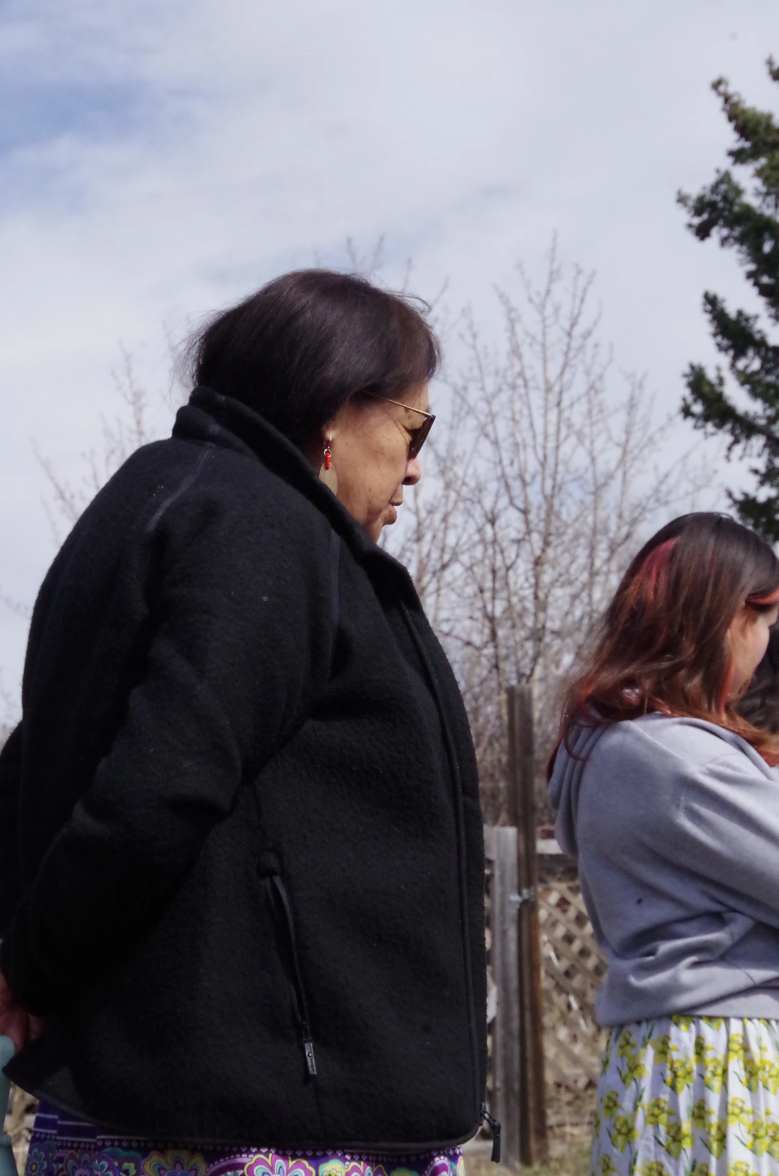 A woman with dark hair, wearing sunglasses, a black jacket, and earrings, standing outdoors with head bowed.