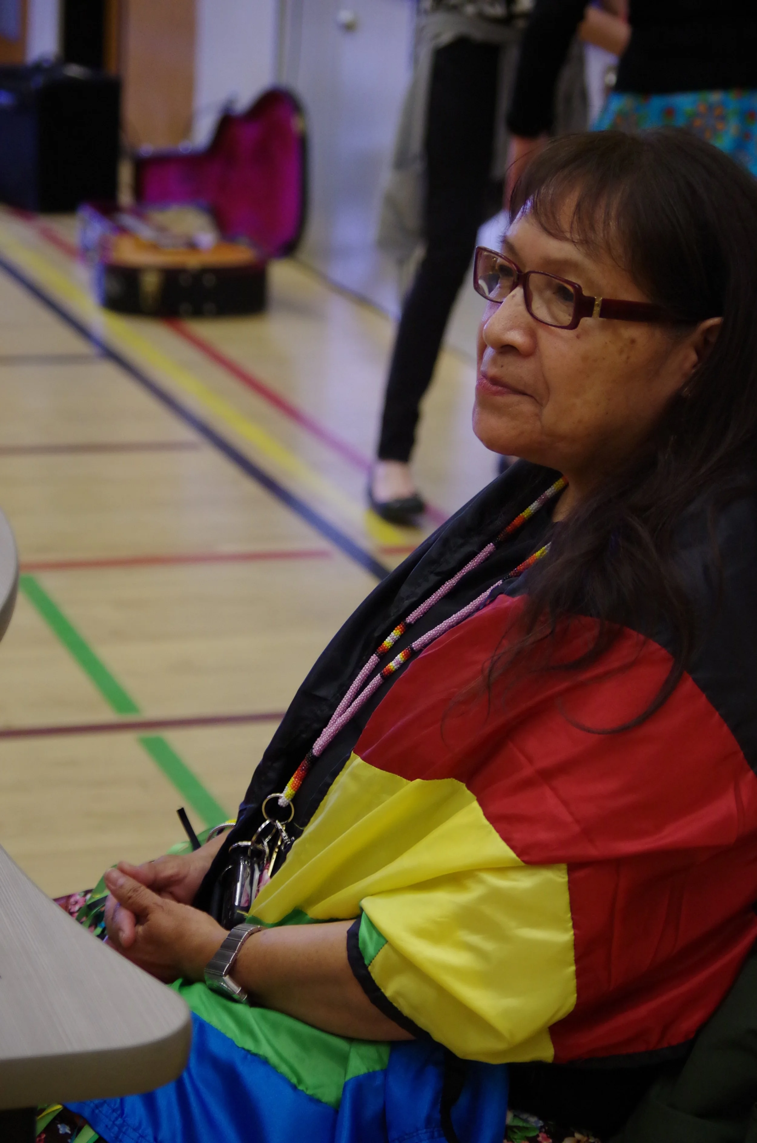 A woman with glasses, wearing a jacket and a colorful necklace, sitting indoors near a table, with a guitar case open in the background.