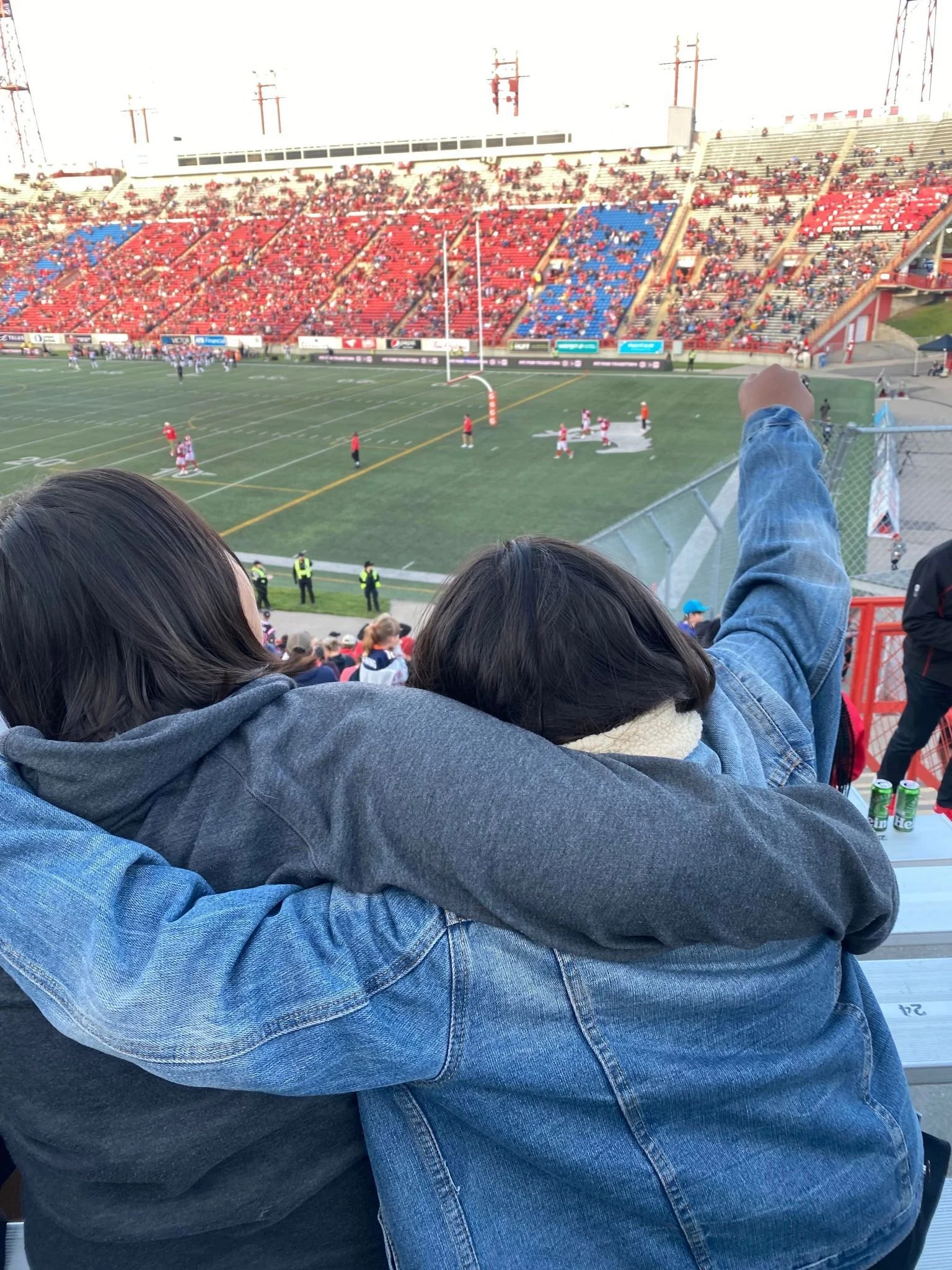 Two friends sitting at a football stadium, watching a game. One is pointing towards the field as the other leans on them, with the field and players visible in the background.