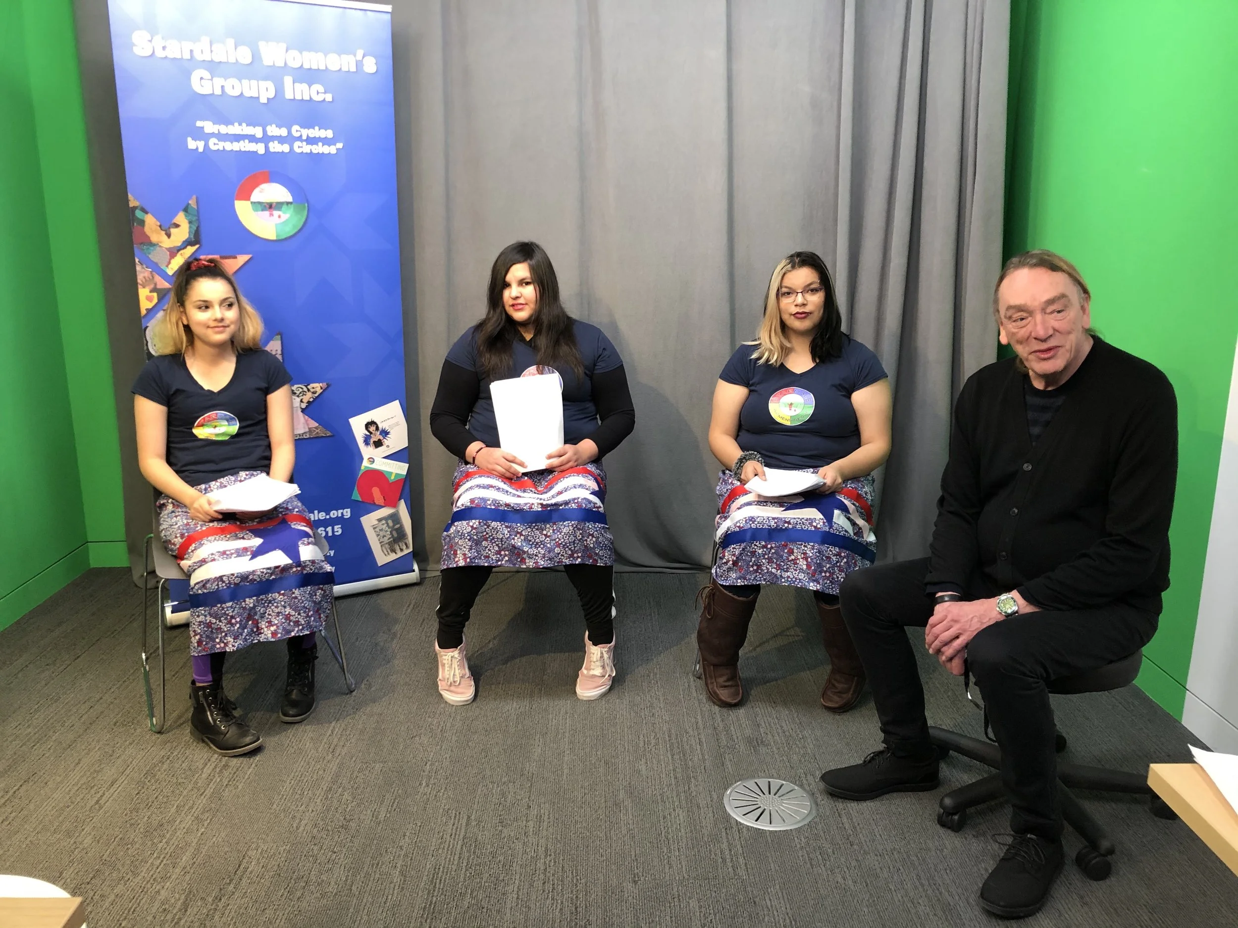 Four women and one man sitting in a row, with a Stanton Women's Group Inc. banner in the background, in a room with green and gray walls.
