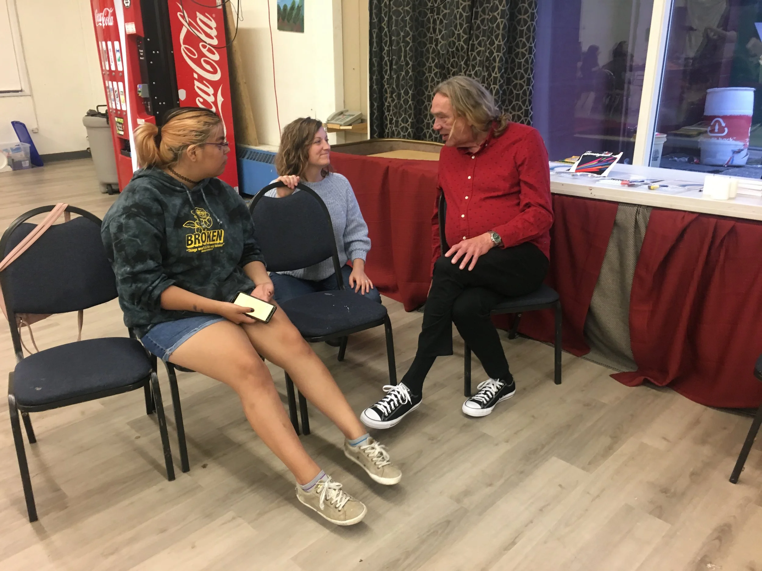 Three people sitting and talking indoors near a red tablecloth and a Coca-Cola vending machine.