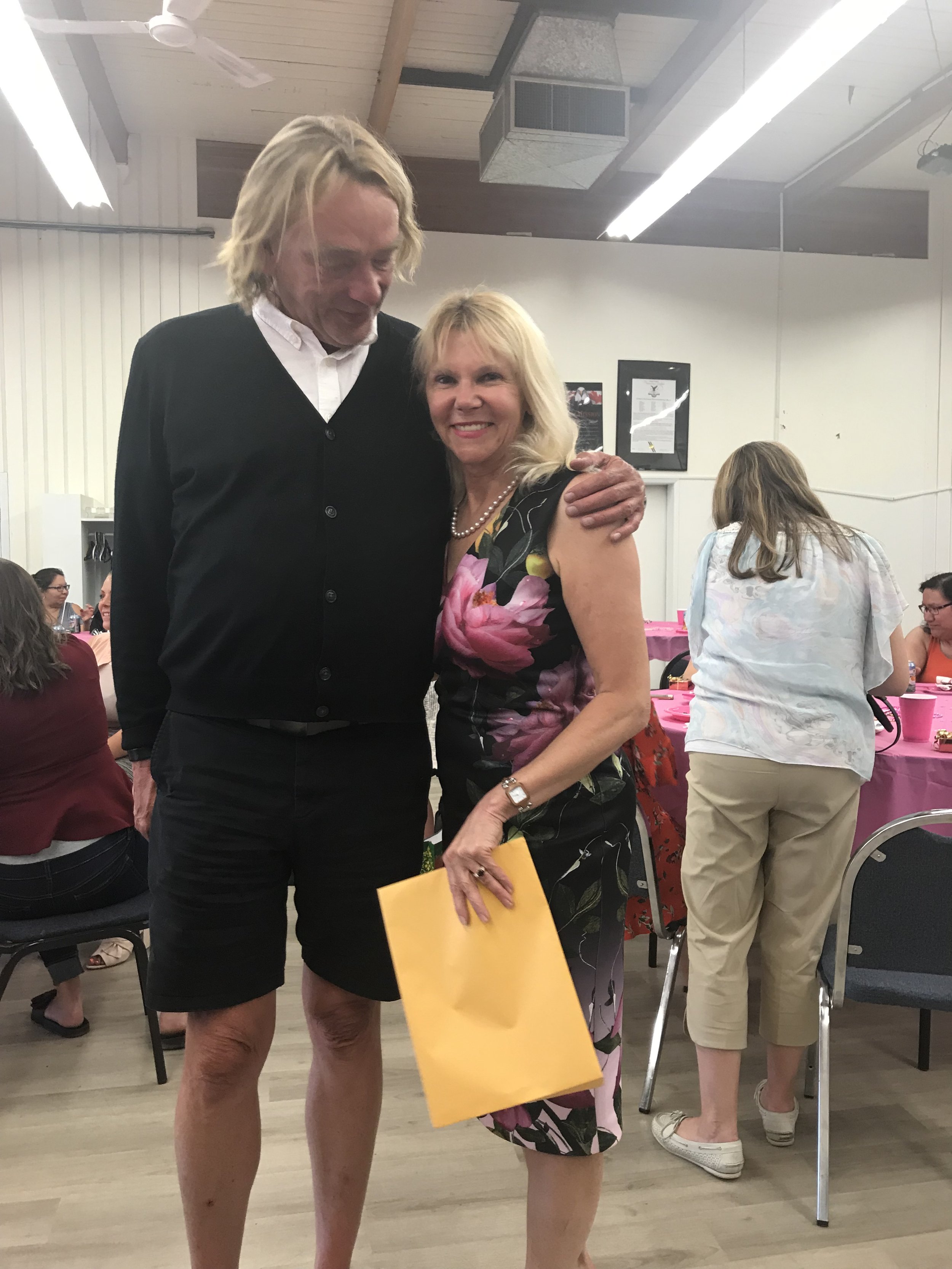 A man and woman are hugging each other, smiling, at a social gathering. The man has blond hair and is wearing a black cardigan over a white shirt, while the woman has blonde hair and is dressed in a floral dress, holding a yellow gift bag.