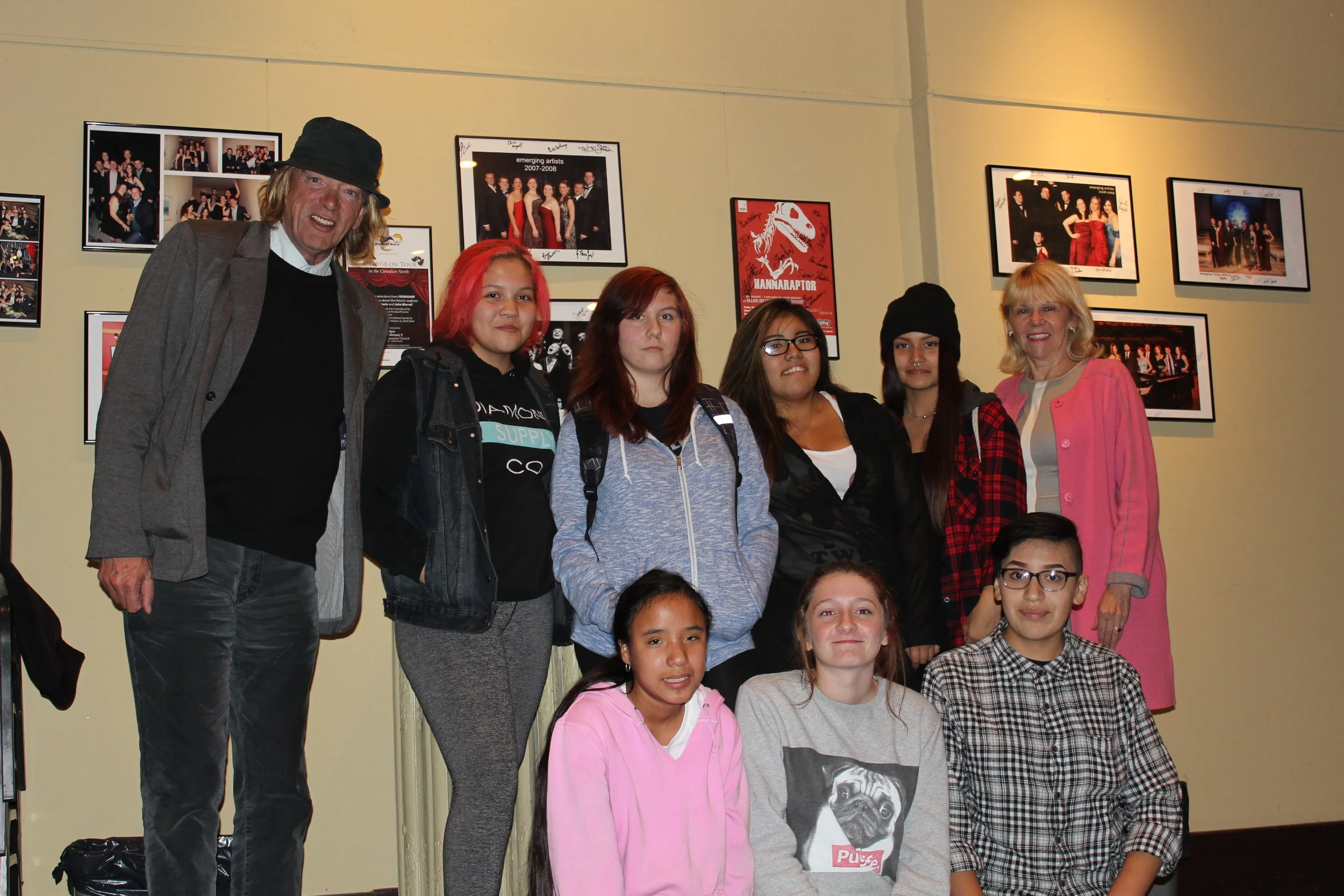 Group of nine people, including two adults and seven children, standing indoors in front of a wall decorated with framed photographs and posters.