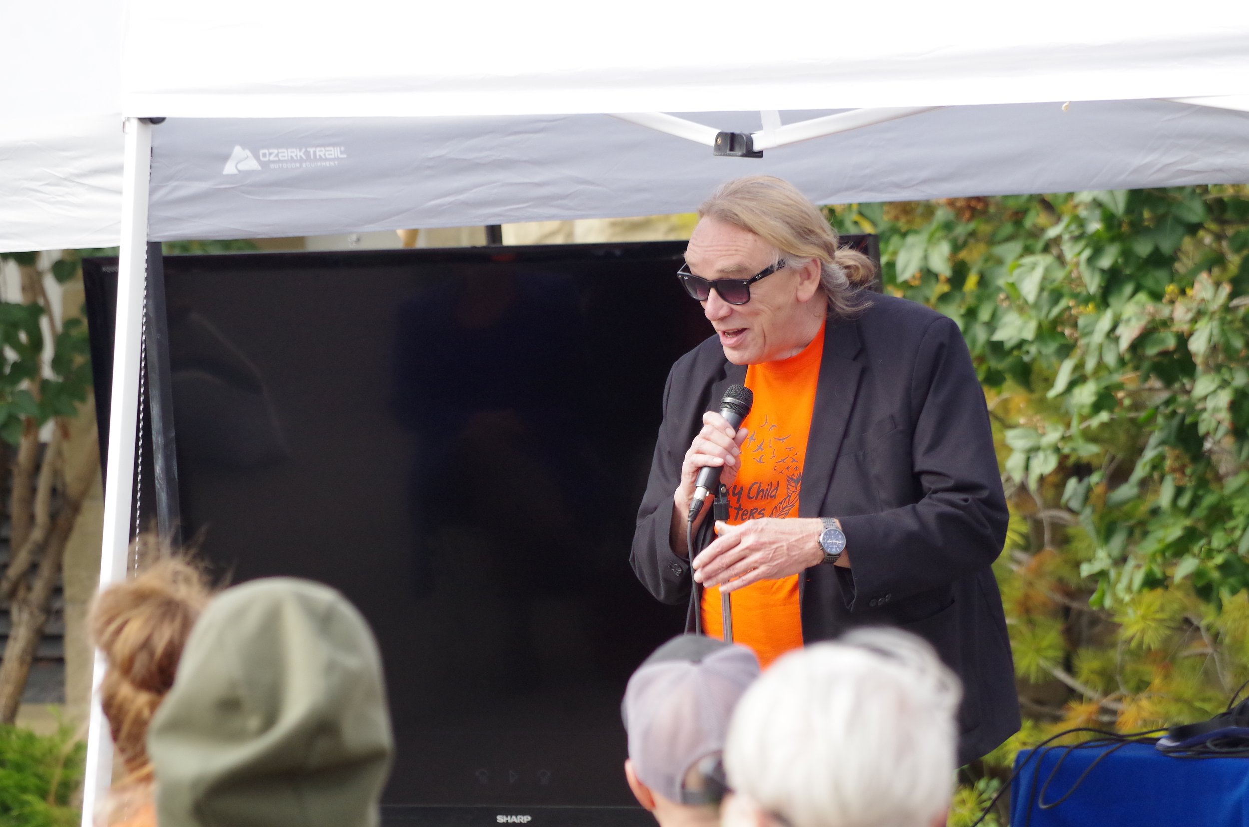 A man with long hair, sunglasses, and a black blazer speaking into a microphone at an outdoor event under a white canopy, with a crowd in front of him and green foliage in the background.