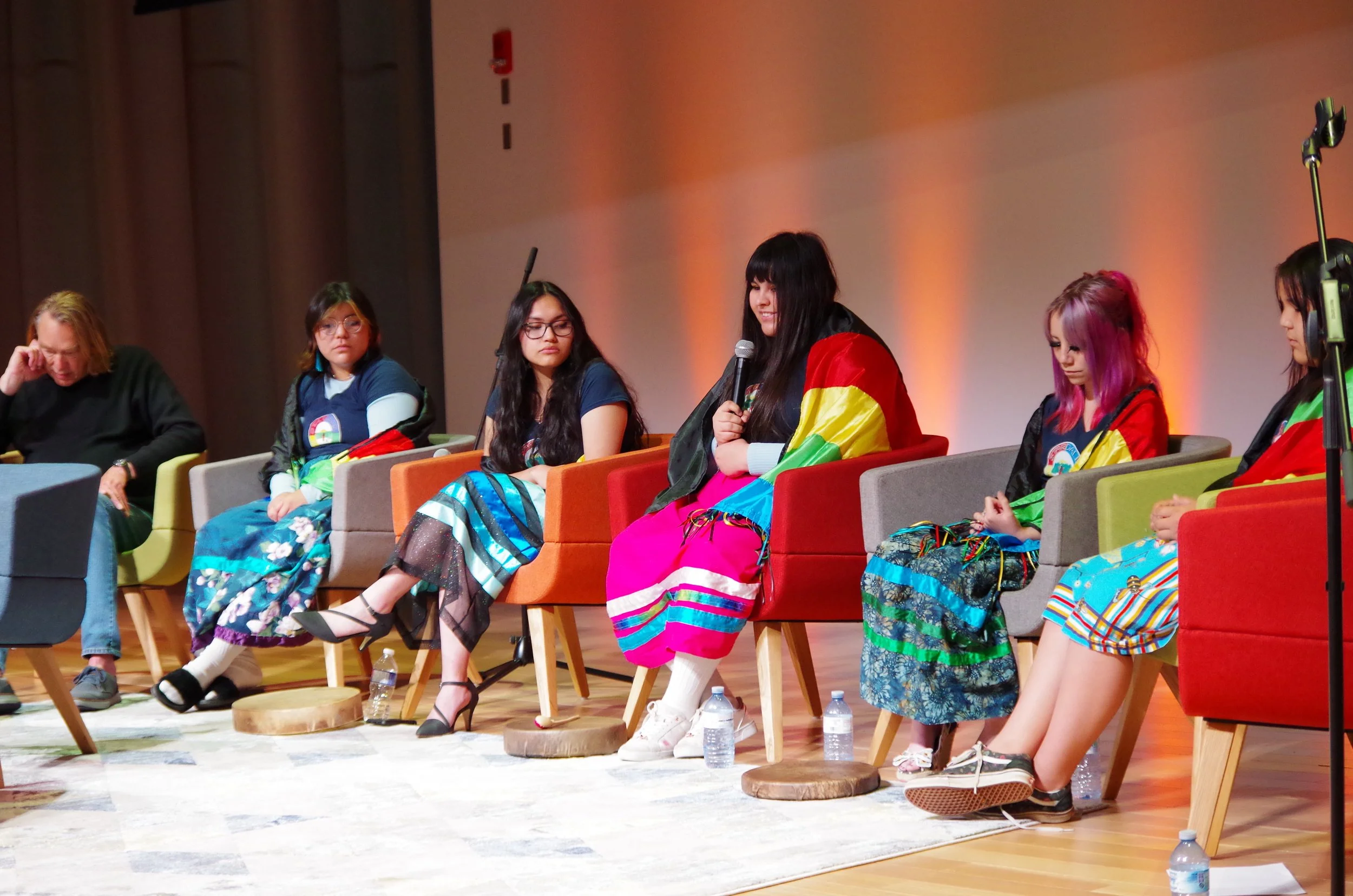 A group of women sitting on chairs on stage, some dressed in colorful traditional clothing, during a panel discussion or event.