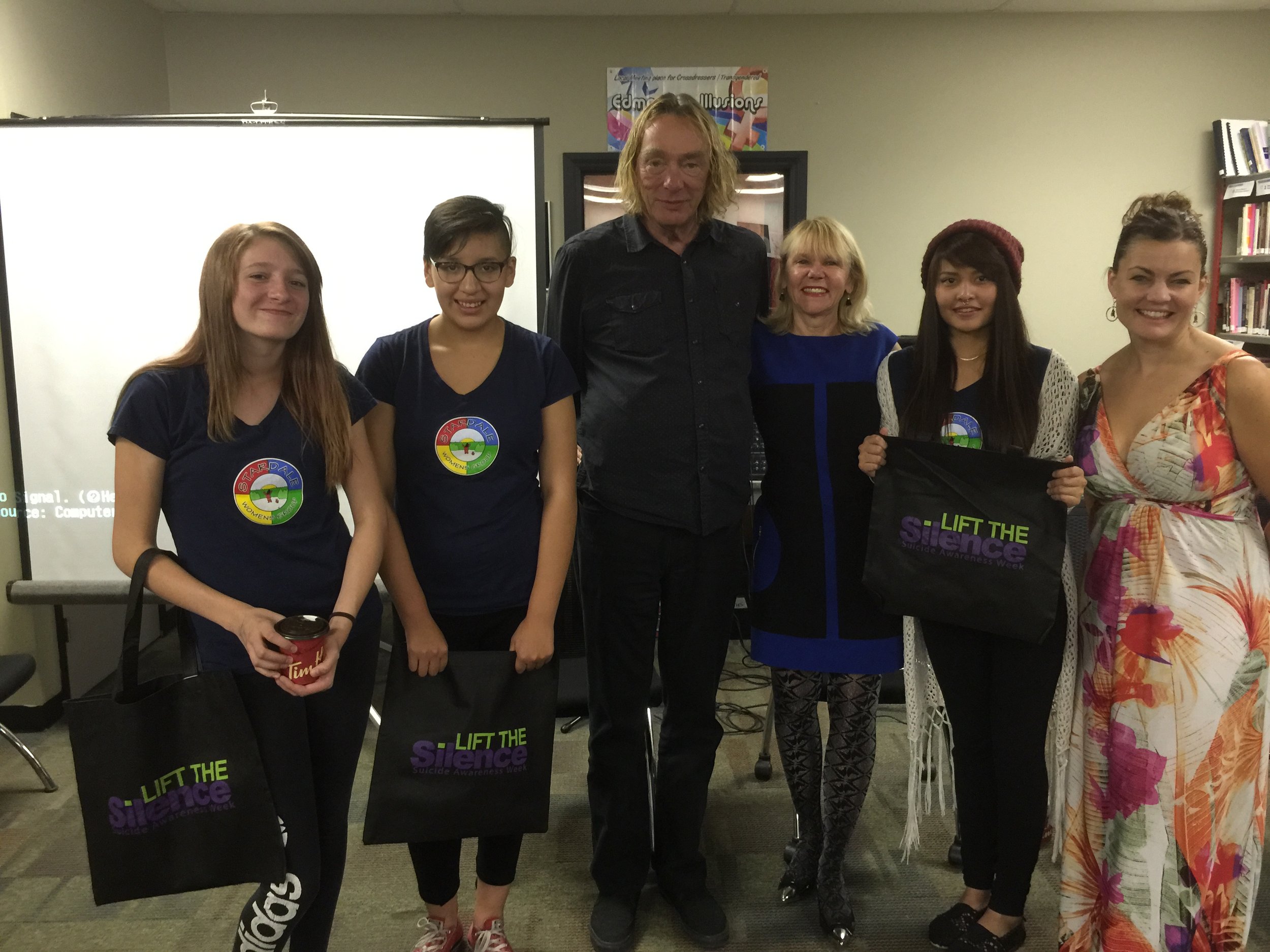 Group of six women and two men standing together indoors at an event, holding tote bags with the text 'Lift the Silence' for Suicide Awareness Week, some women are wearing navy shirts with a colorful logo.