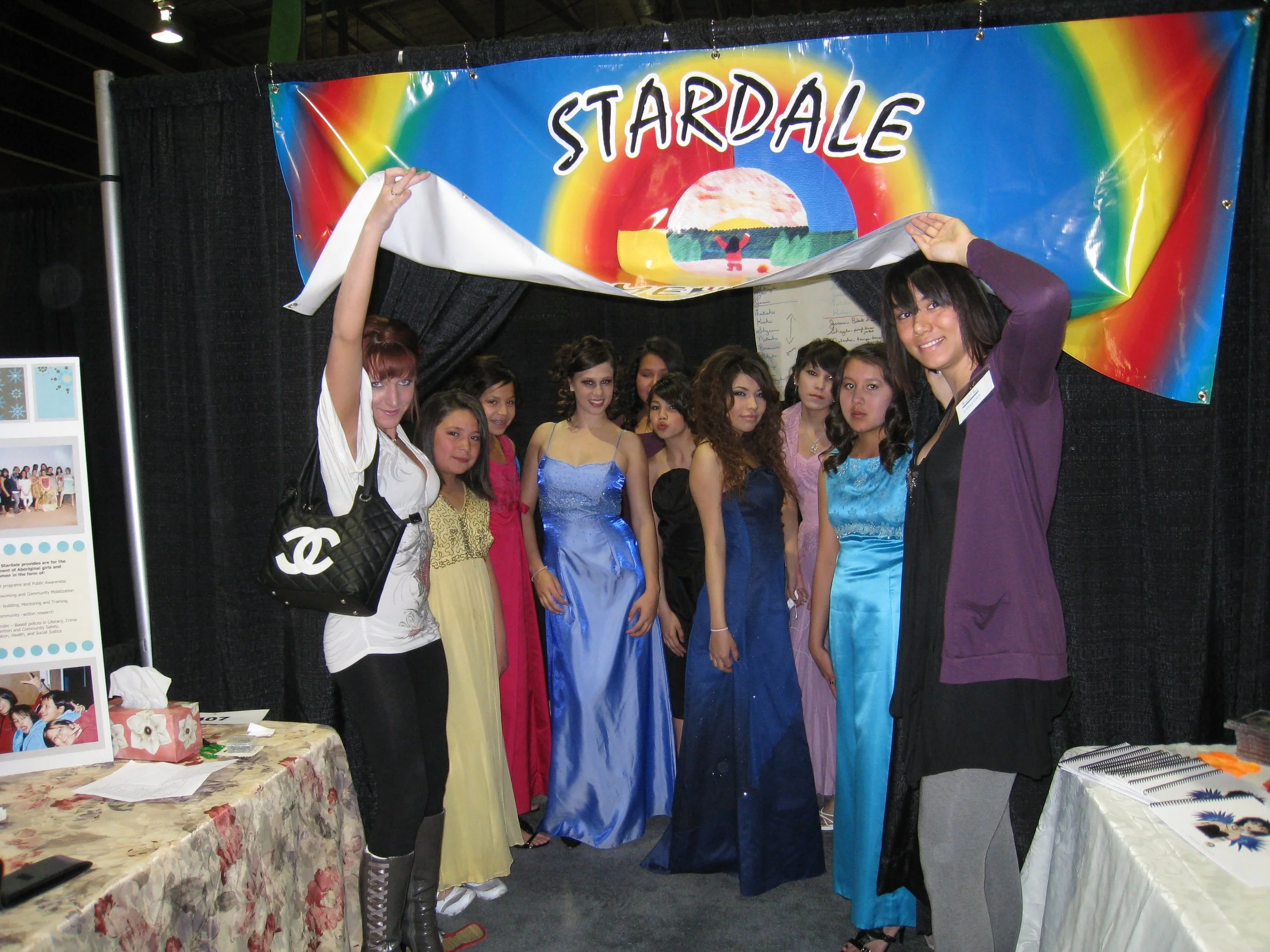 Group of young women standing in front of a colorful banner that says 'STARDAL'. Some women are wearing formal dresses, and one woman on the right is lifting the banner with a woman on the left holding it up. There are tables with sponsor information and materials on either side.