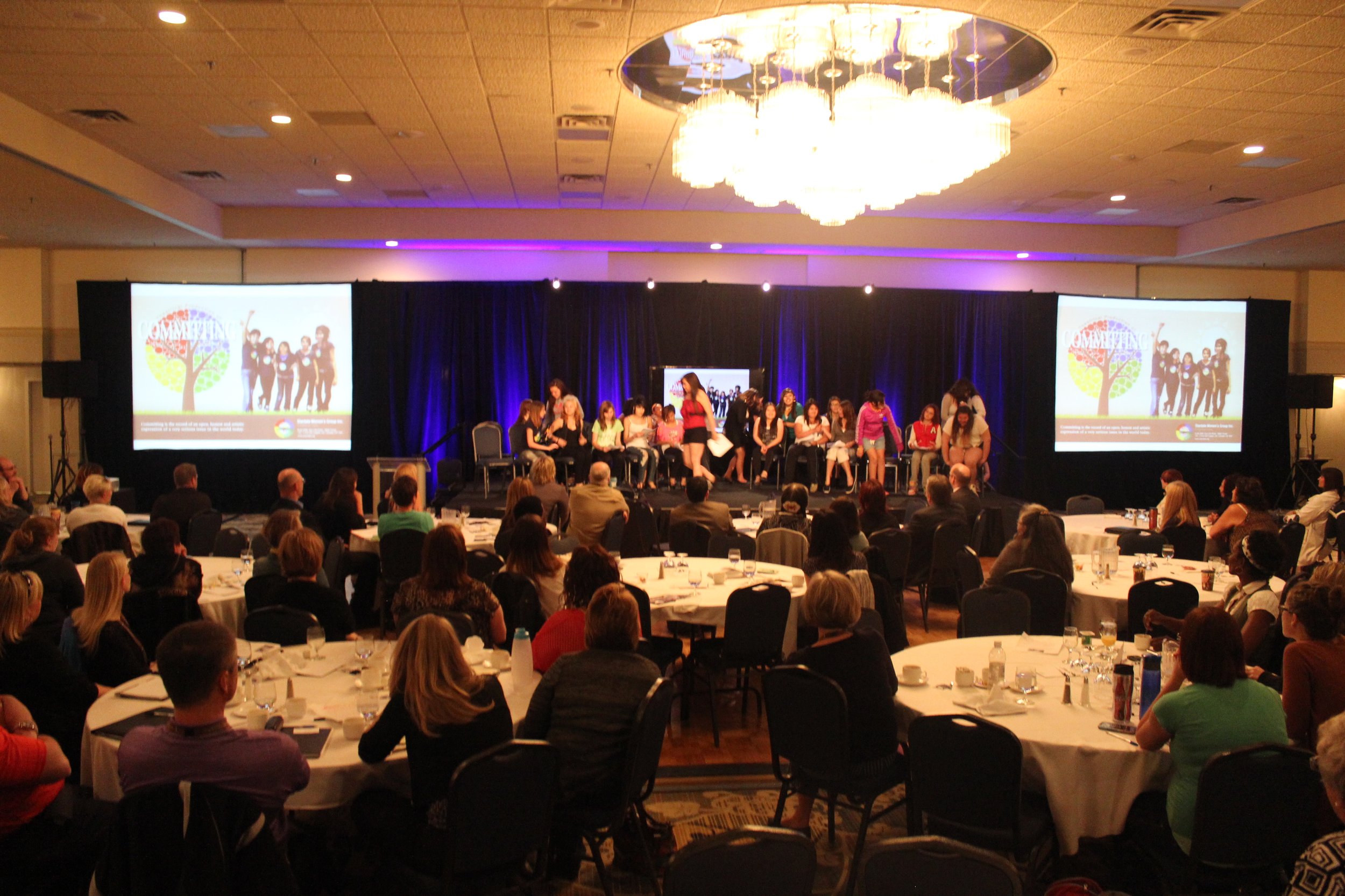 A large conference room filled with round tables and attendees facing a stage. On the stage, a group of people sits while a woman in a pink top stands speaking. Two large screens on either side display a colorful logo with a tree and the word 'COMMUNITY' along with images of people. The room is illuminated with warm lighting, and a large chandelier hangs from the ceiling.