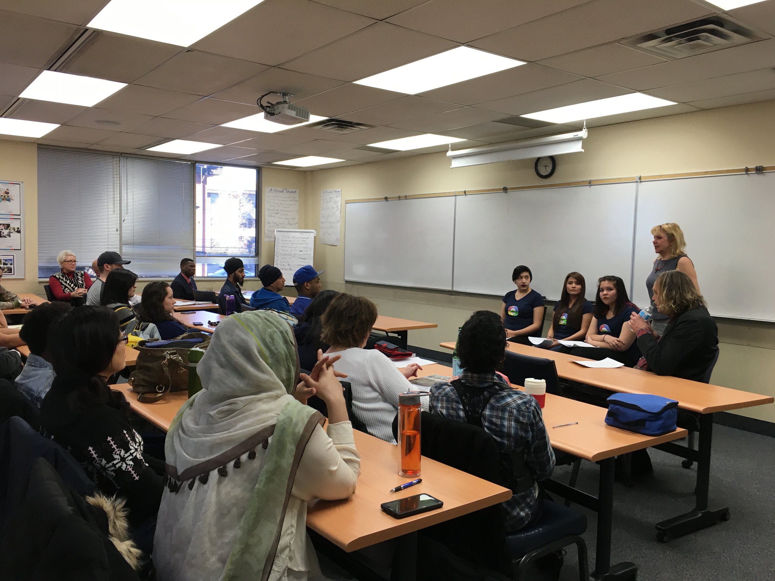 A classroom with students sitting at tables, facing a teacher and panel of four women at the front, with a whiteboard and clock on the wall.