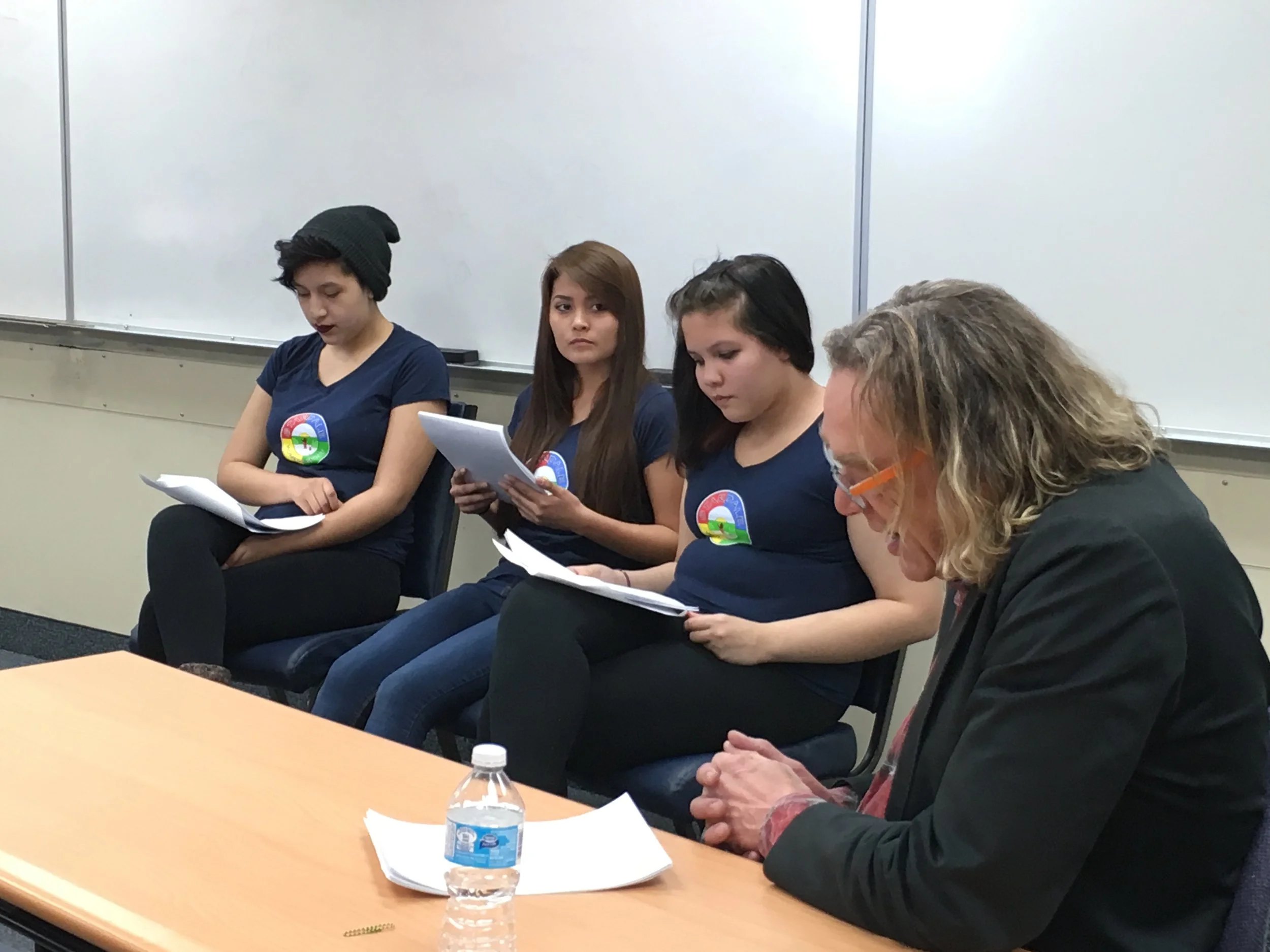 Four women sitting in a row, looking at papers, in a classroom with a whiteboard behind them. One woman has a water bottle and papers on the table in front of her.