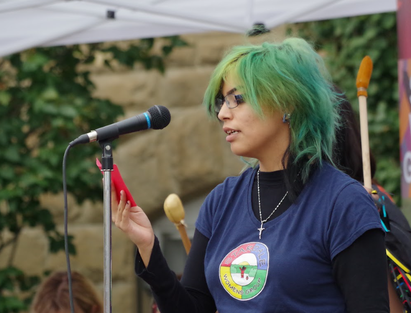 A young woman with green and blue hair speaking into a microphone at an outdoor event, wearing glasses and a navy blue T-shirt with a logo that reads 'Starlake Women's Group.' She is holding a red phone or device and wears a silver cross necklace. There are trees and a white canopy in the background.