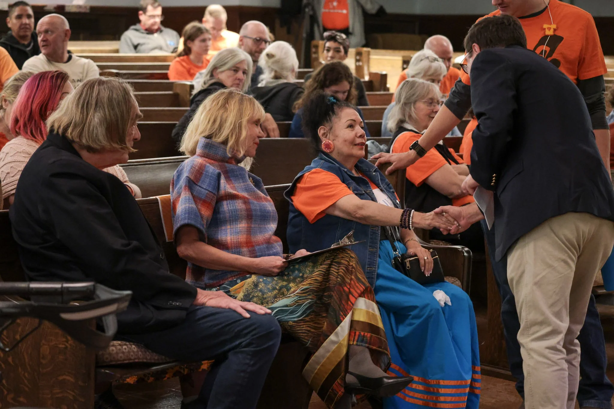 A woman in an orange shirt and denim vest shaking hands with a man in a dark blazer in a room with wooden pews and other seated attendees, some of whom are dressed in orange shirts.
