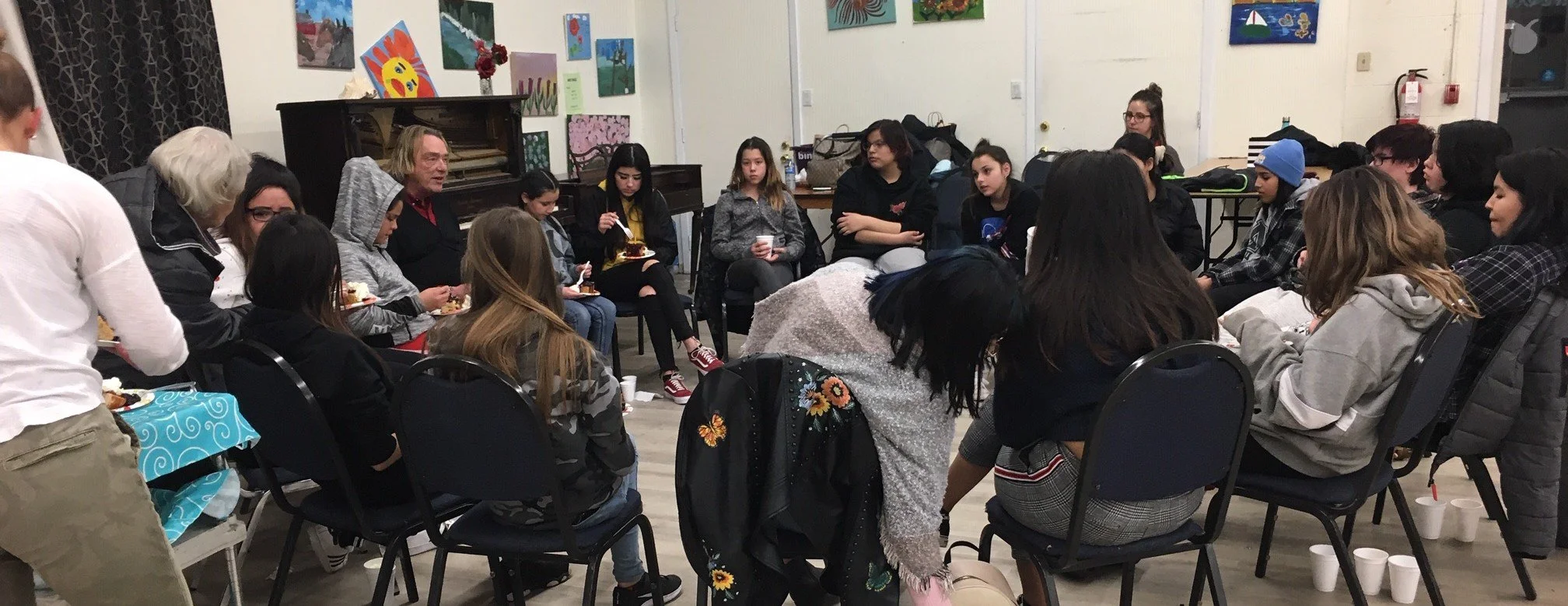 A group of people, mostly young women and girls, sitting in a circle in a room decorated with colorful paintings, engaged in a social gathering or discussion, with some holding food and drinks.