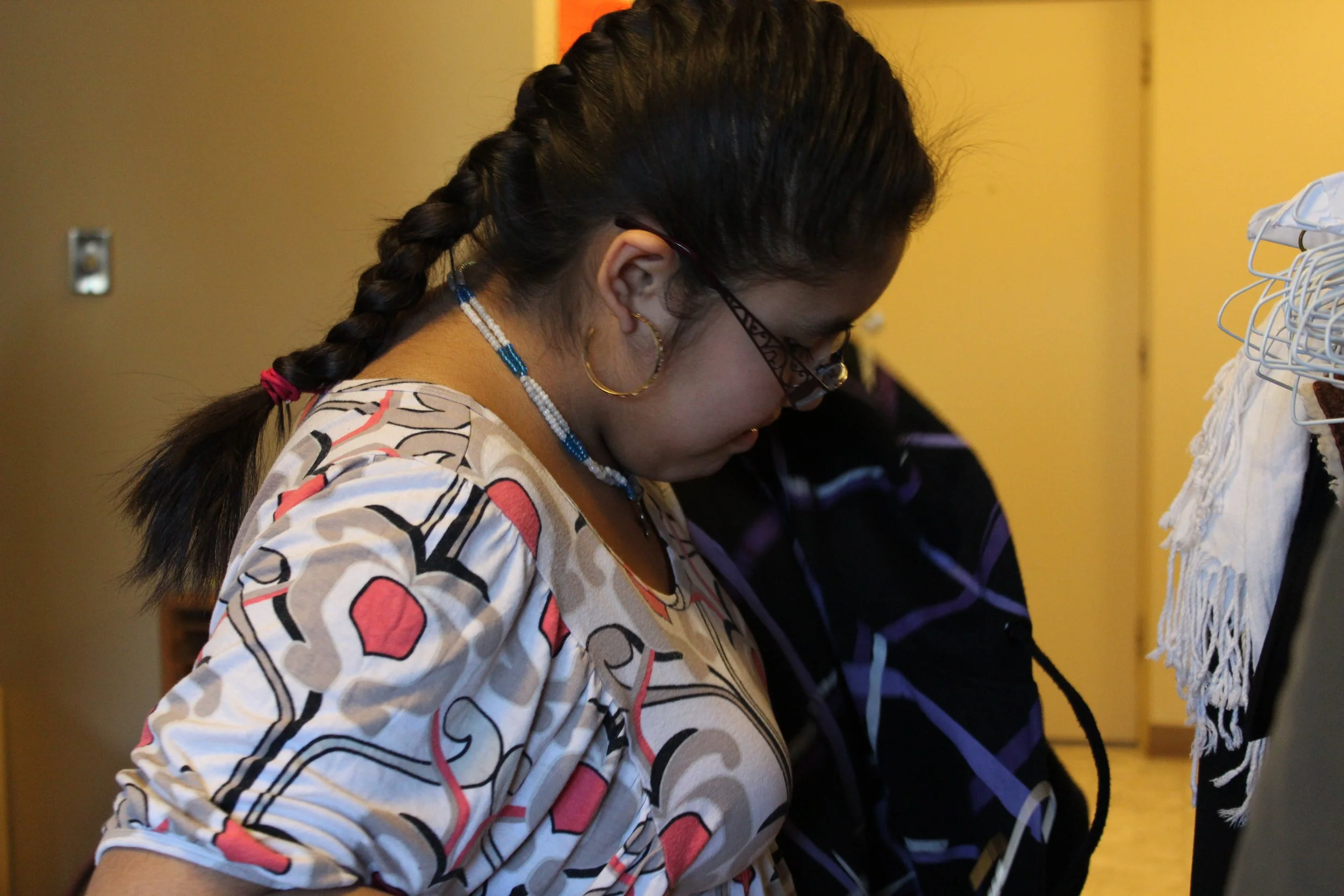 A woman with glasses, hoop earrings, and a beaded necklace looking down, wearing a colorful patterned top, with her hair in a braid, standing next to a rack of clothing in a room with yellow walls.