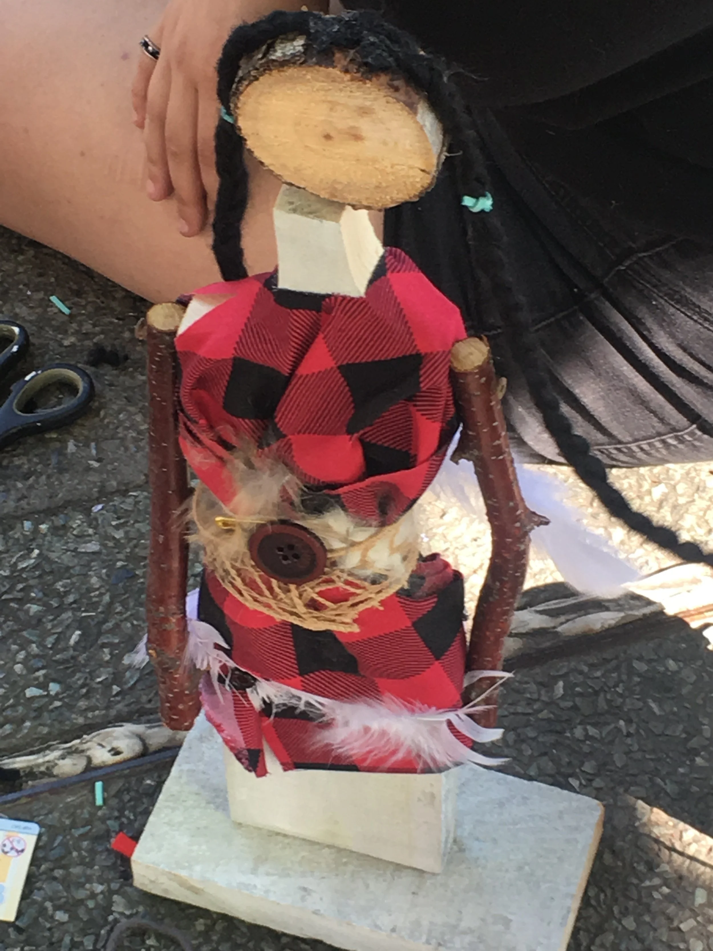 A handmade doll with a wooden head, fabric body, and small twigs for arms, dressed in a red and black checkered fabric with a button, placed on a stone base.