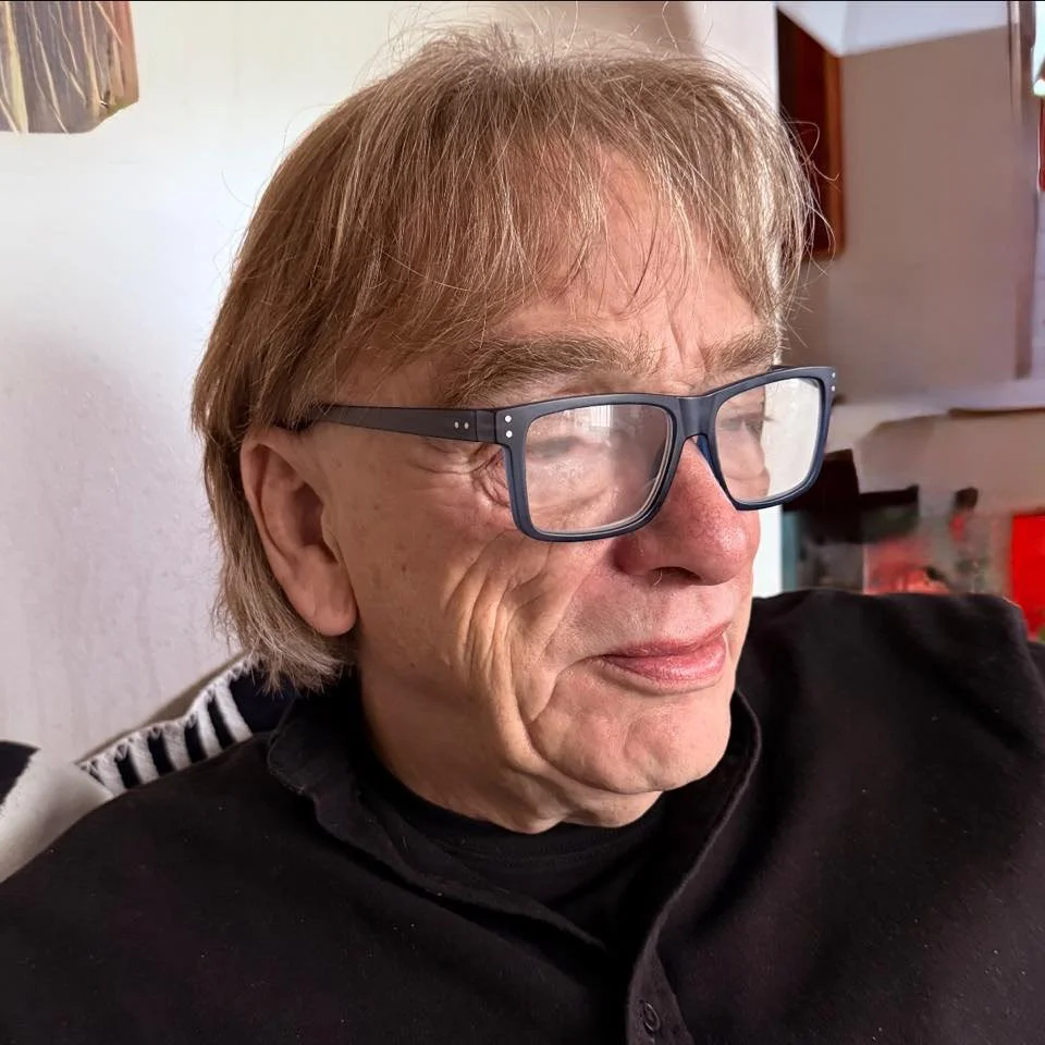 Close-up of an elderly man with glasses, light brown hair, and wearing a black shirt, sitting indoors.
