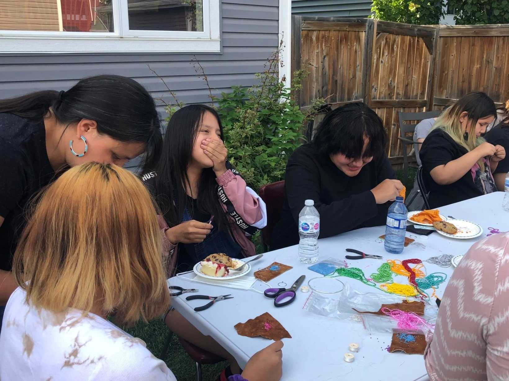 People sitting at a table outdoors, engaged in a crafting activity with colorful yarn and supplies, surrounded by a wooden fence and greenery.