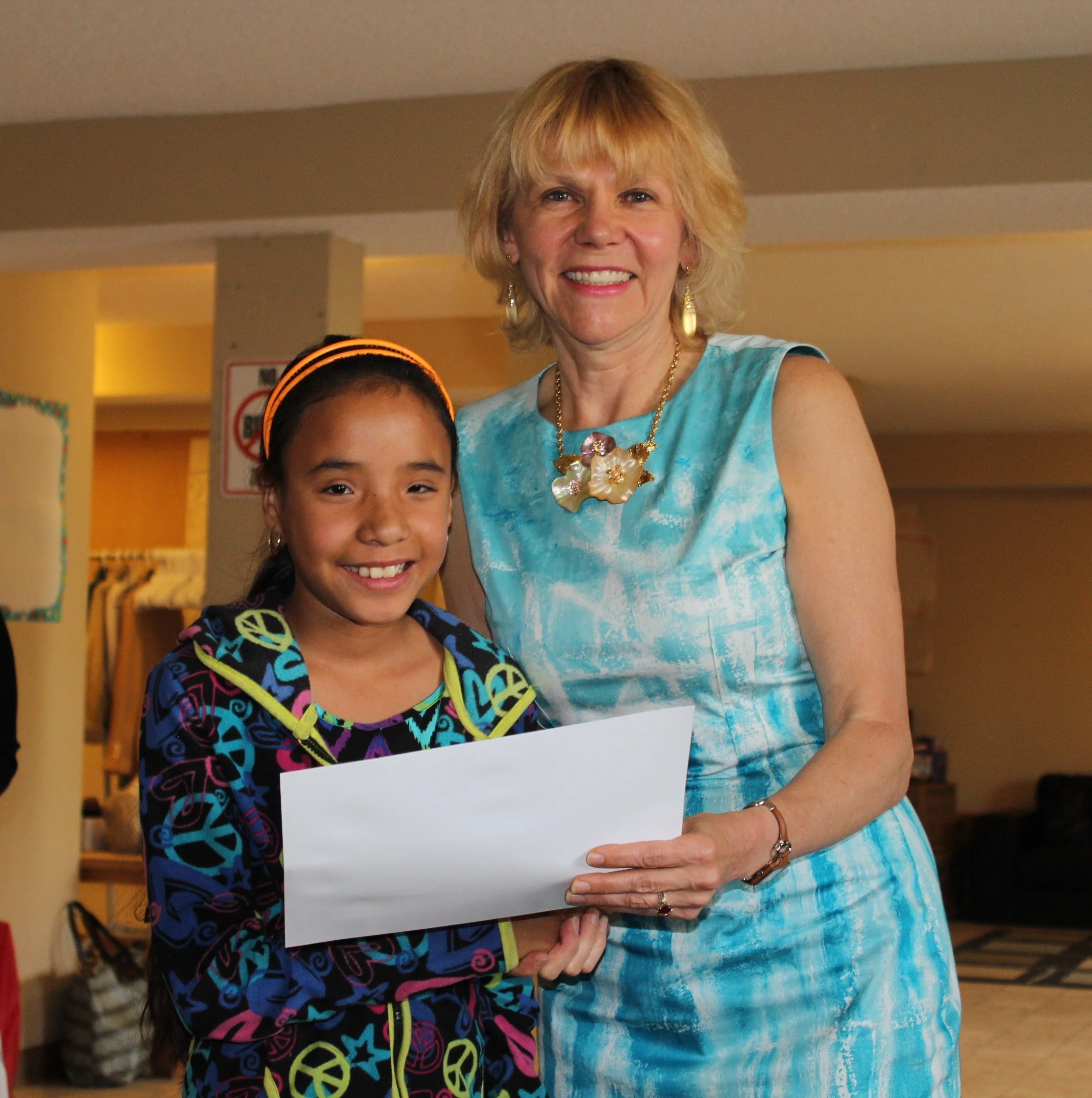 A young girl with dark hair, wearing colorful peace sign patterned hoodie, holding a white envelope, standing next to an older woman in a light blue dress with floral jewelry, in an indoor setting.