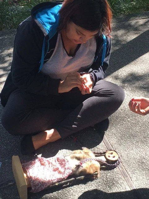 A woman crouches on a sidewalk, looking down at a small animal lying on a makeshift table. The animal appears to be a rat with some injuries, wrapped in a cloth. The woman holds a small object in her hands.