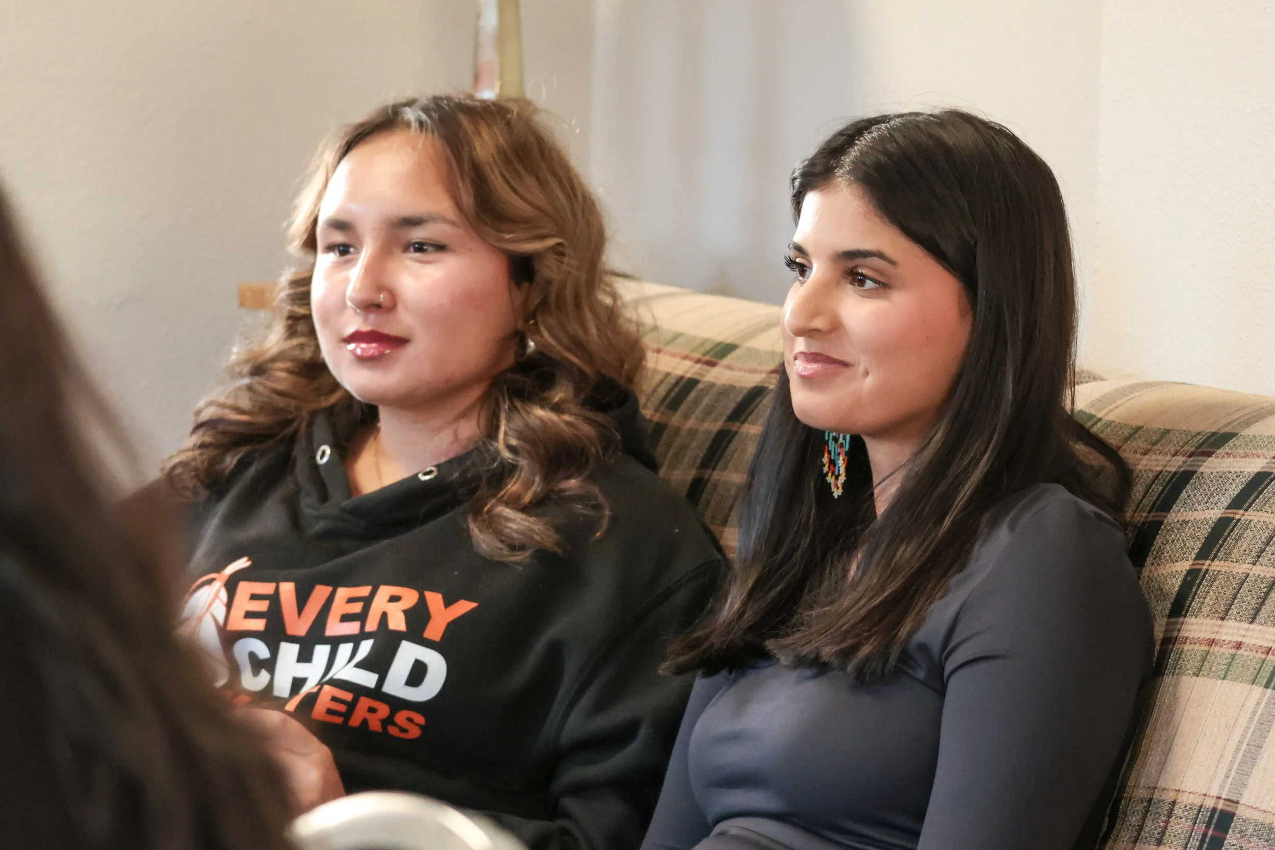 Two women sitting on a plaid couch, engaged in conversation, in a cozy room with beige walls.