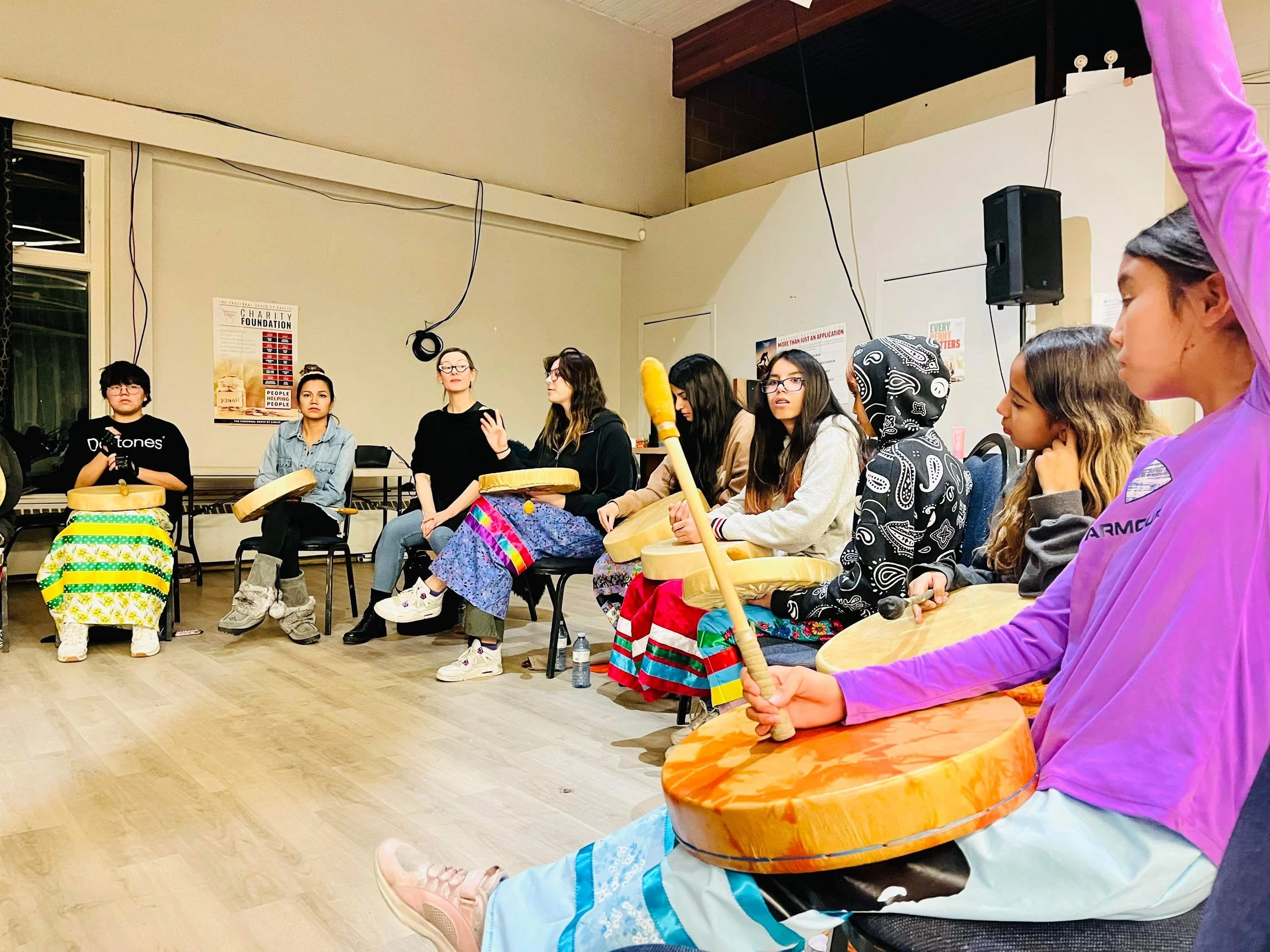 A group of women sitting in a circle, some holding drums and some holding microphones, participating in a drumming circle or workshop in a room with posters on the wall.