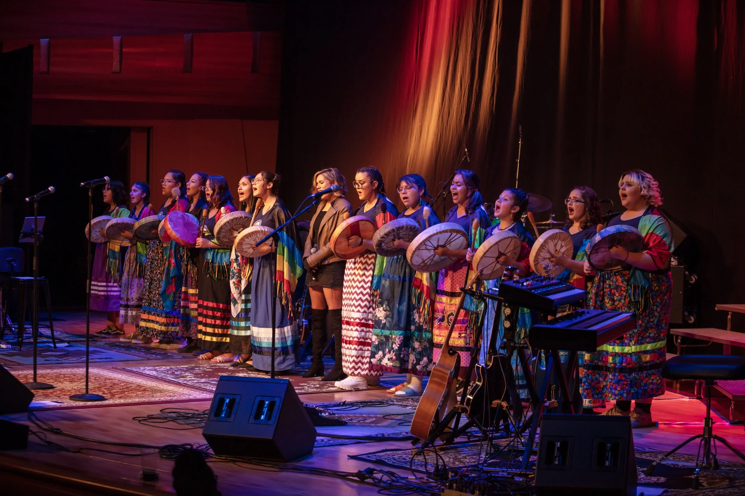 A group of women performing on stage, singing and holding traditional Asian fans, dressed in colorful traditional attire, with musical instruments and microphones around them.