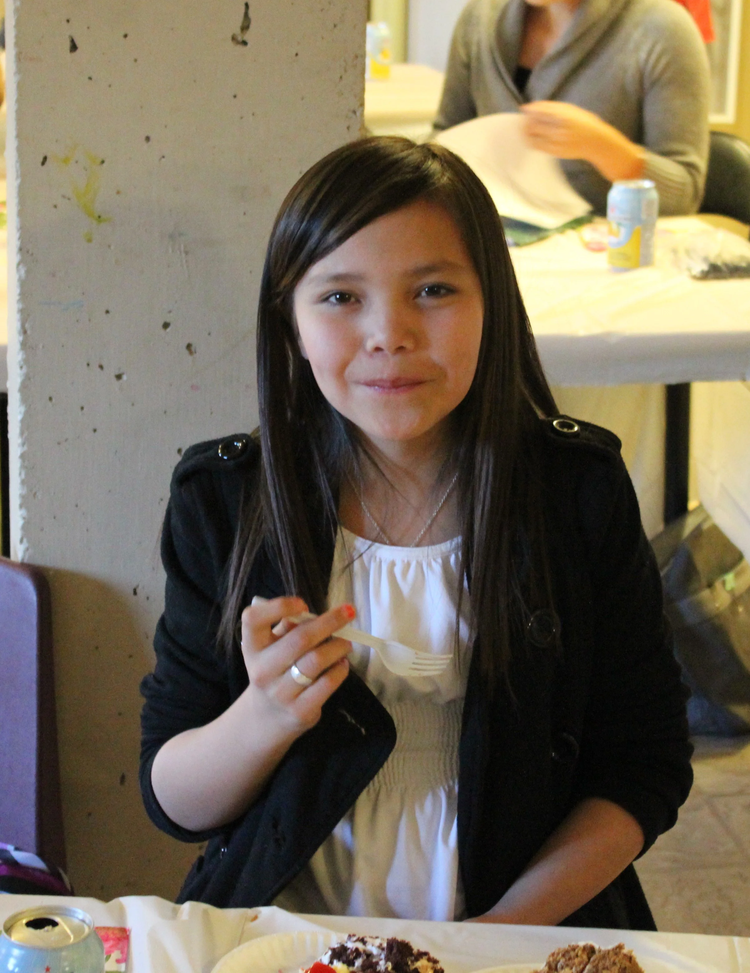 A young girl with long dark hair and a black jacket, holding a fork, sitting at a table with cake and desserts, smiling at the camera.