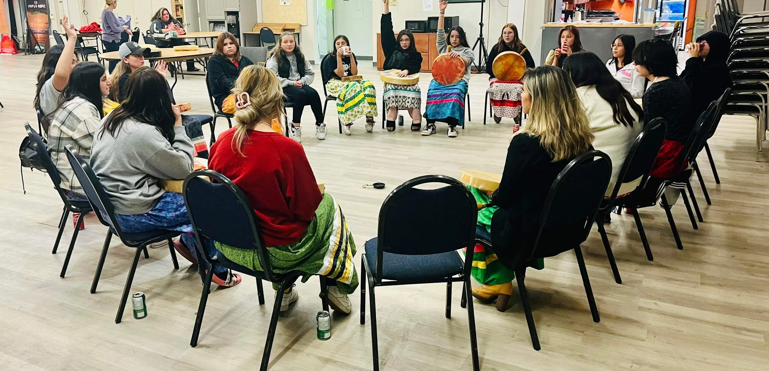 Group of women sitting in a circle, some raising hands, in a community center or classroom setting with chairs, tables, and various items around.