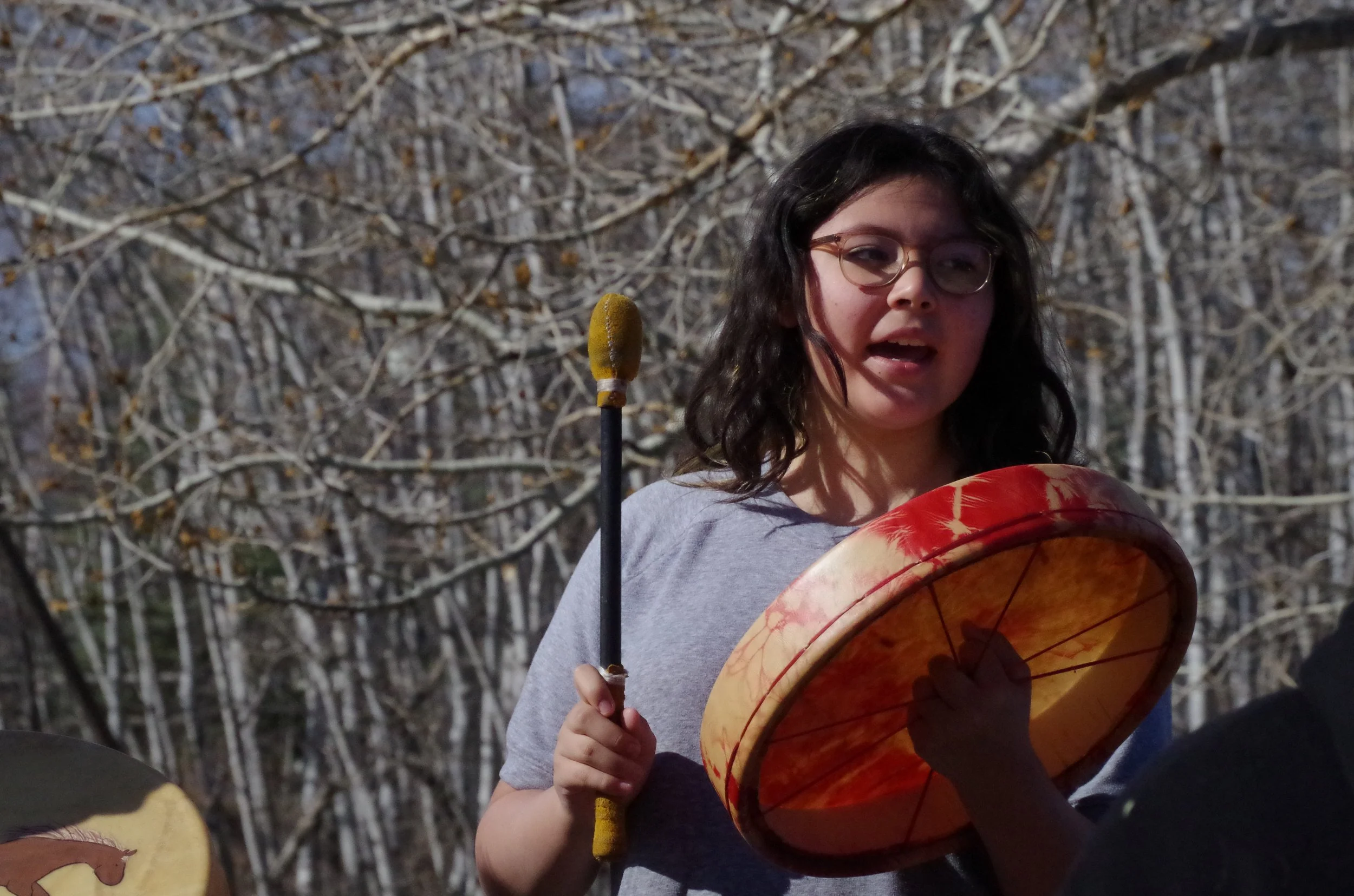 A young woman with glasses playing a drum outdoors, with leafless trees in the background.