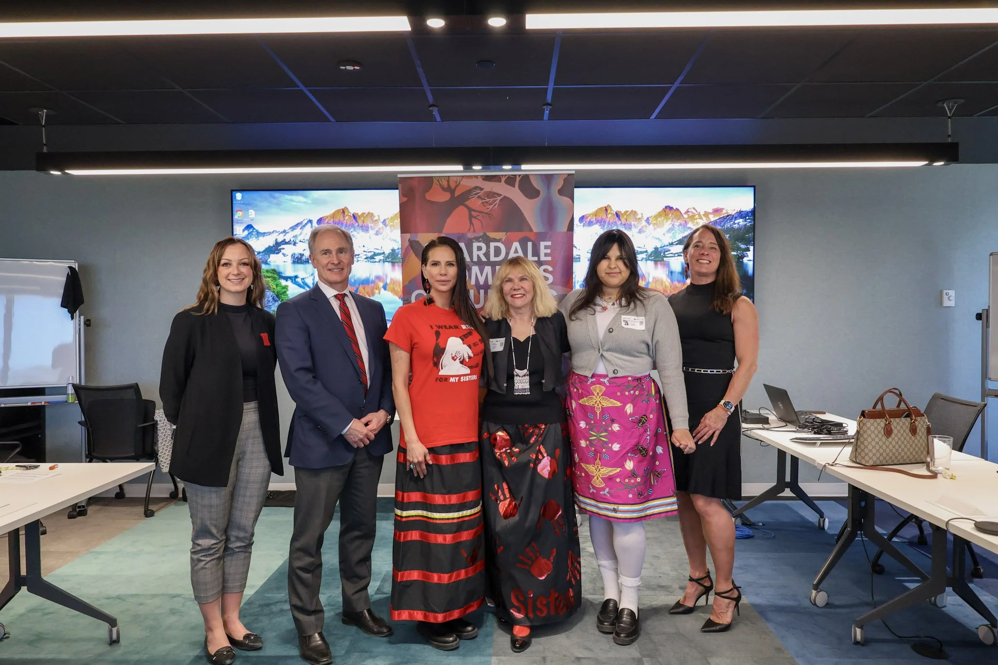 Six women and one man standing together in a conference room, smiling for a photo, with a banner reading "HARDALE WOMENS' SUMMIT" behind them, and two large screens displaying mountain scenery in the background.