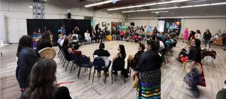 People gathered in a large indoor space, some sitting in chairs and others standing, with various handmade craft items in the background.