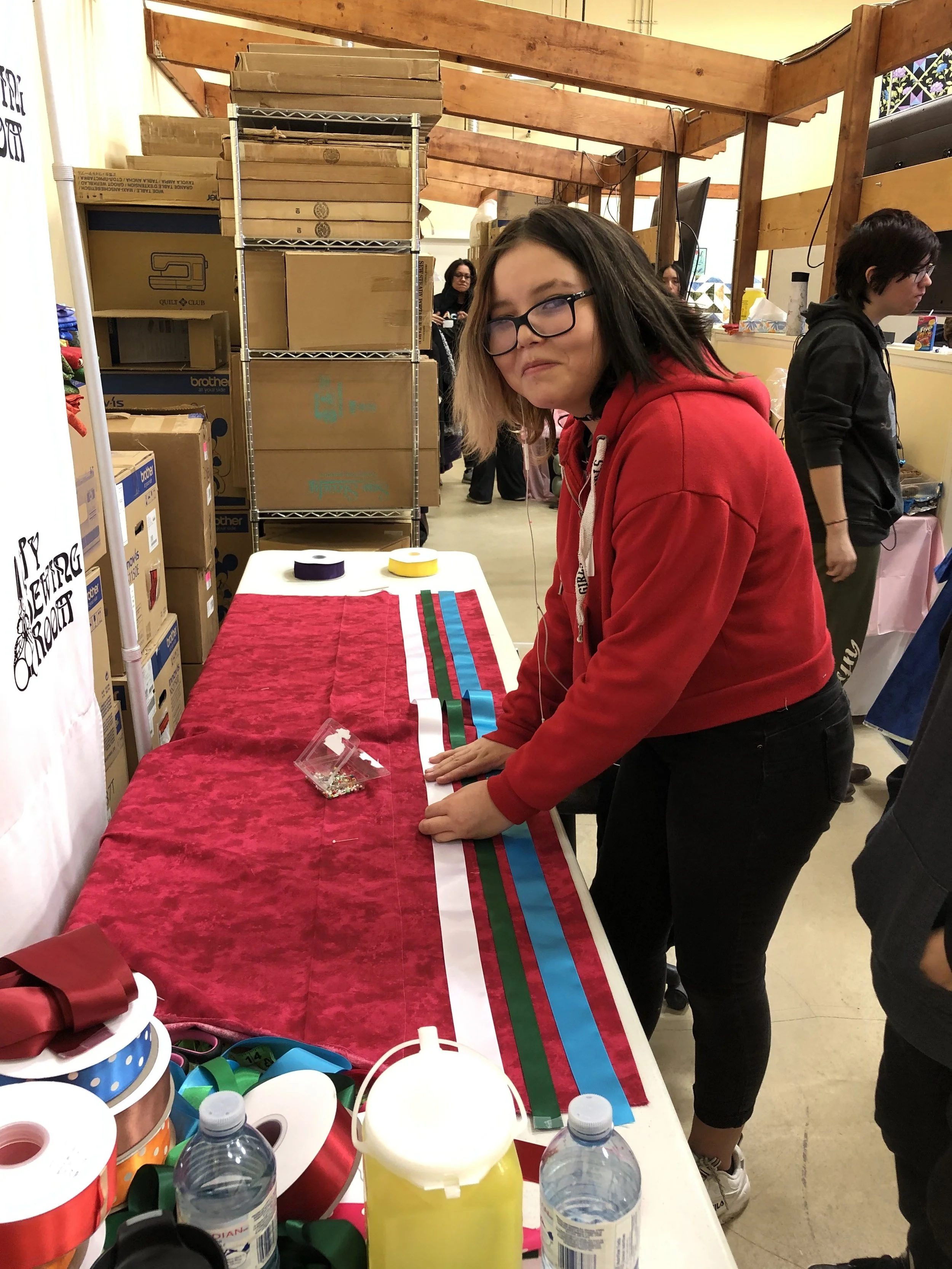 A young woman with glasses and a red hoodie is standing at a table covering with red fabric and rainbow-colored ribbons, preparing for a craft project. Behind her are cardboard boxes, metal shelves, and other people in the background at indoor craft 