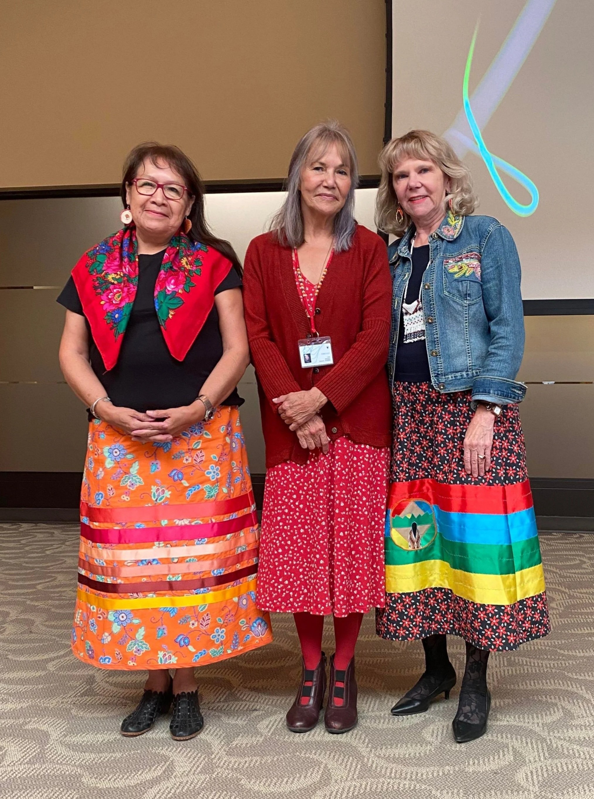 Three women standing side by side indoors, dressed in colorful clothing with floral patterns, in front of a beige and black wall with a projector screen.