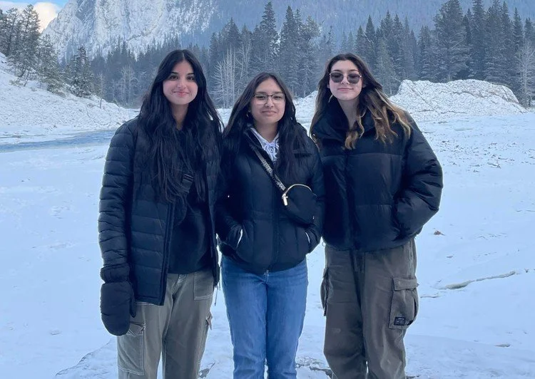 Three young women standing outdoors in a snowy landscape with mountains and trees in the background.