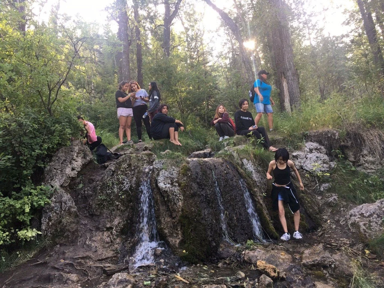Group of people sitting and standing on rocks near a small waterfall in a forest, with sunlight filtering through trees.