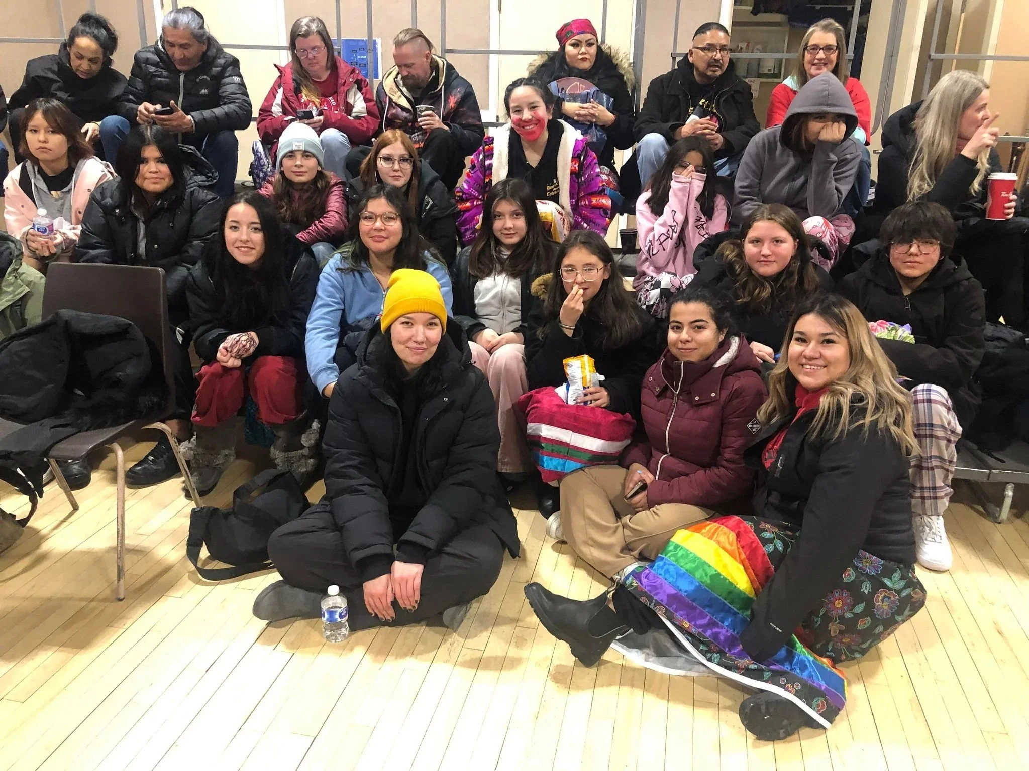 A group of people, including children and adults, sitting together in an indoor space, some wearing winter clothing and holding drinks and snacks.