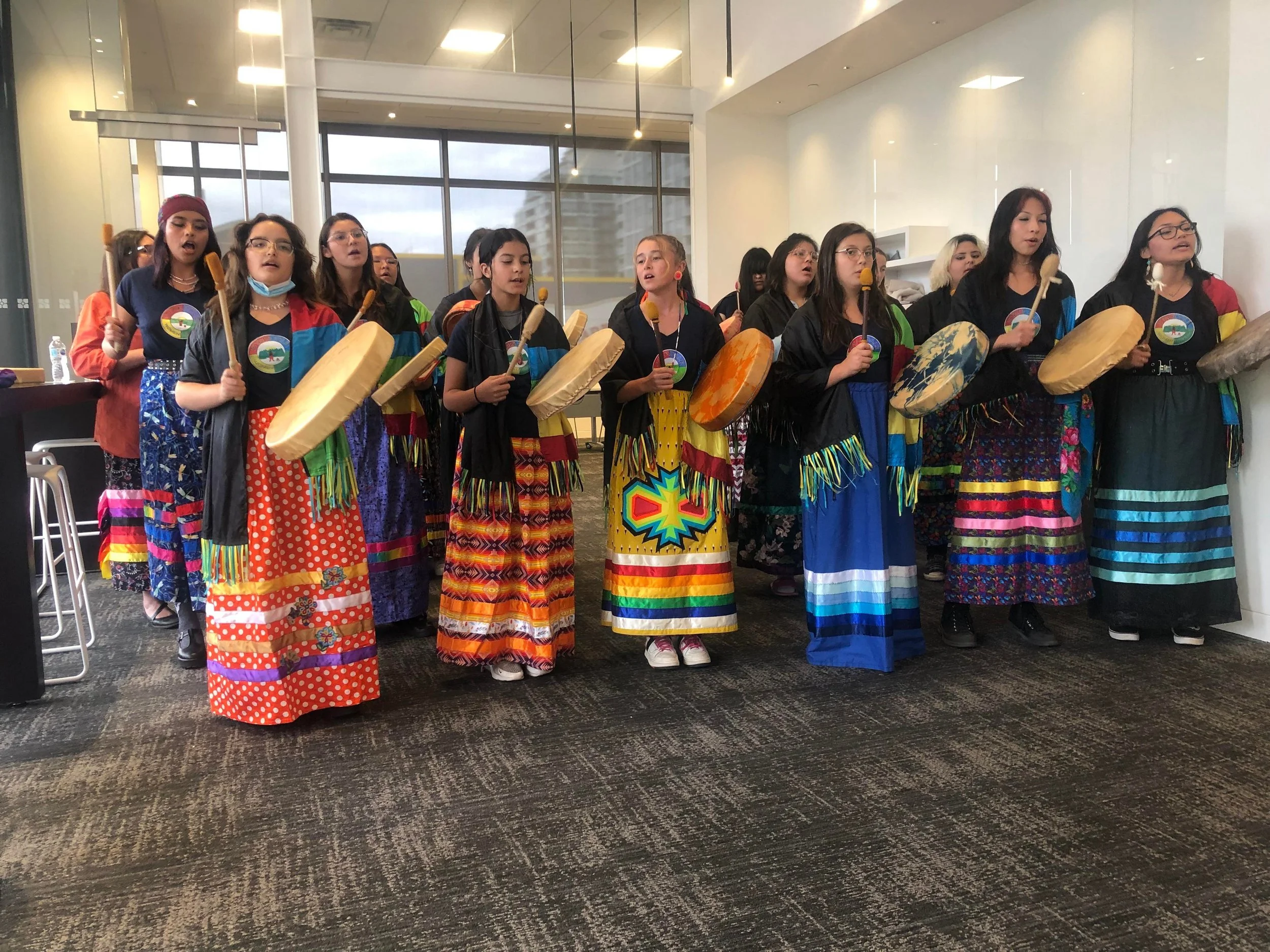 Group of young girls in colorful traditional attire playing drums during a cultural performance indoors.
