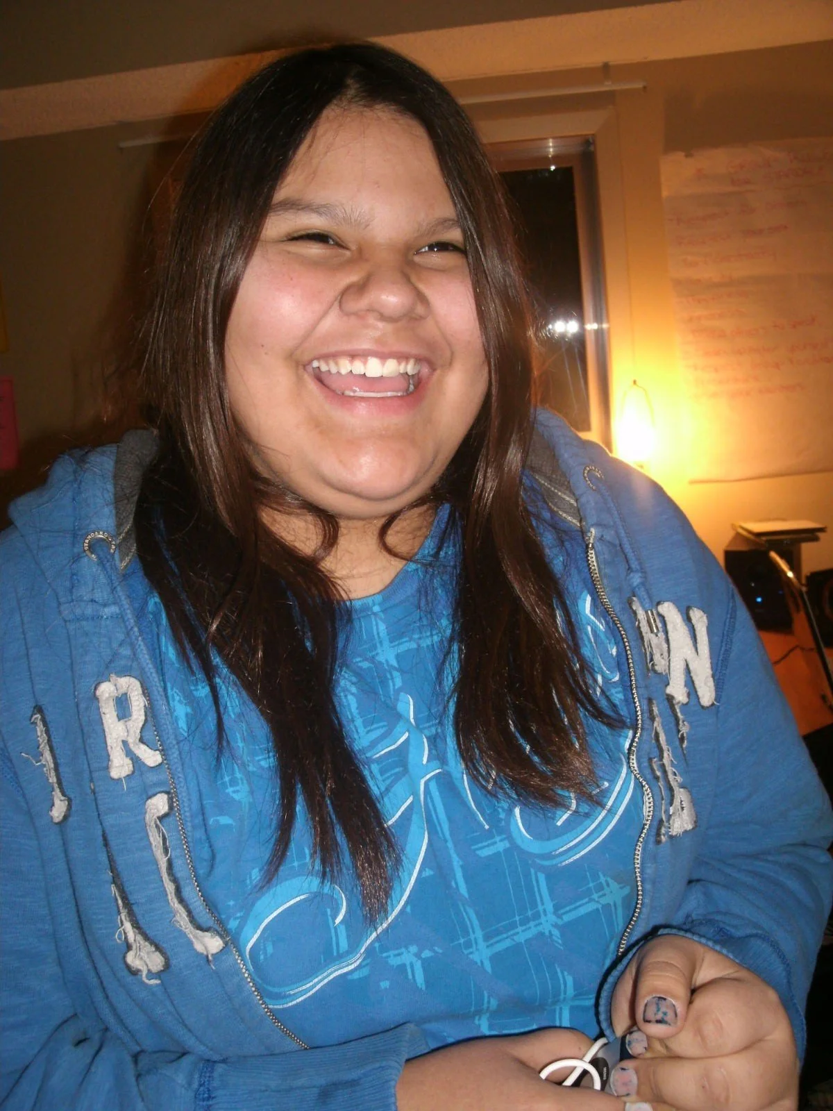 A woman smiling broadly showing her teeth, with long dark hair, wearing a blue hoodie with white lettering, indoors with a background that includes a wall with writing and a warm light.
