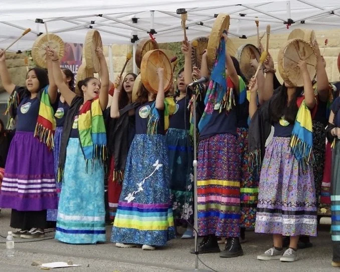A group of young girls dressed in traditional Mexican clothing performing a folkloric dance, holding sombreros in the air, under a white tent at an outdoor event.