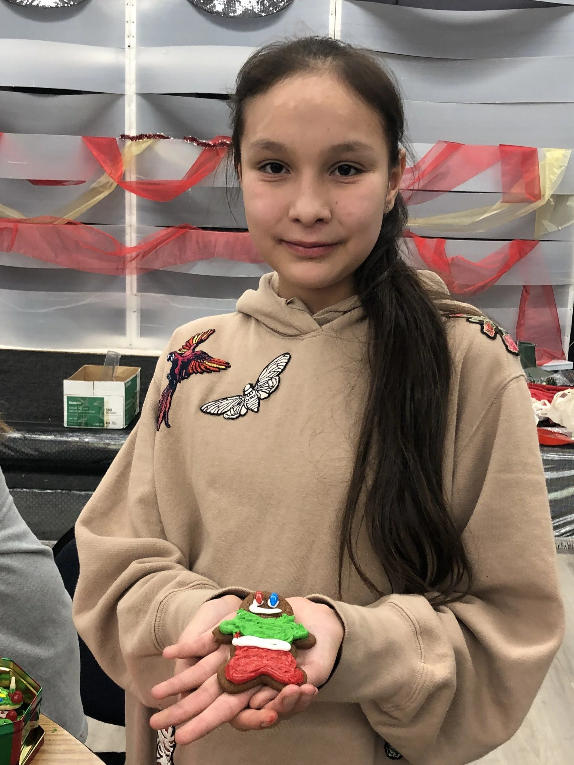 A girl with long dark hair holding decorated gingerbread cookie shaped like a Christmas elf. She is wearing a beige hoodie with patches of a bird, butterfly, and flowers. The background has gray wall panels with red and yellow streamers.